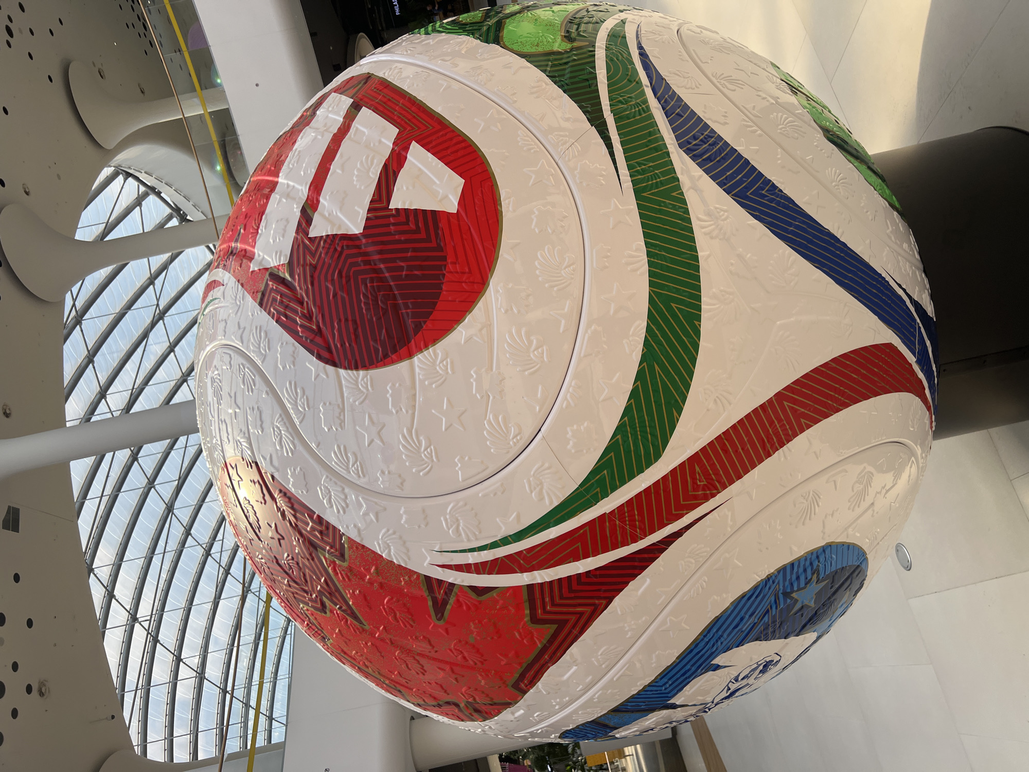 The image shows a large, spherical sculpture resembling a soccer ball, decorated with various colors and patterns. The ball is predominantly white with red, green, blue, and gold accents. The white sections are embossed with intricate designs of leaves and stars. Red and white sections on the top appear to form a flag-like emblem, with red stripes and white bars. Colored bands of red, green, and blue with gold outlining wrap around the ball. The sculpture is positioned indoors, likely in a public space with a modern architectural design, characterized by a curved, transparent ceiling allowing natural light to filter in, supported by white pillars. The flooring is light-colored and smooth. The location is identified as Kyiv, Ukraine. No people are clearly visible in the immediate foreground, but faint outlines suggest the presence of others in the background.