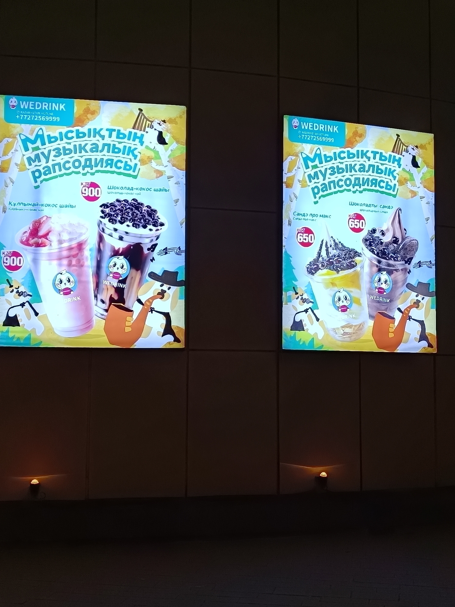 The image shows two illuminated advertisements for a brand called "Wedrink," featuring stylized cartoon characters and drinks, likely displayed on a wall in a public area, perhaps in the city of Kokshetau, Kazakhstan (given the presence of Kazakh language text and a Kazakhstan hotline number). The ads promote beverages with flavors such as strawberry-coconut, chocolate-coconut, and "Sande pro max" alongside chocolate sundaes. Prices in KZT are displayed.