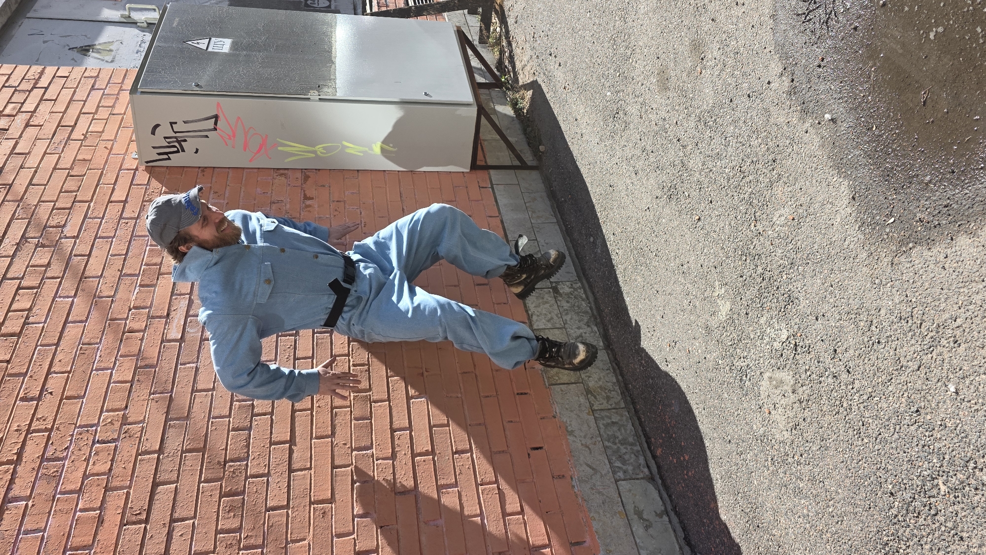 The image displays an optical illusion photograph of a man posing in an urban outdoor environment in Saint Petersburg, Russia. The photo is rotated 90 degrees clockwise, making the man appear to be standing vertically on a brick wall, with a utility box to his side and an asphalt surface below.
In its true orientation, the man is lying on his side on an asphalt ground, leaning against a reddish-orange brick wall. He is a bearded individual wearing a light blue denim-like jumpsuit, a grey baseball cap, and rugged dark brown boots. He exhibits a subtle smile, indicative of a playful pose for the trick photography. His arms are slightly bent and hands raised, contributing to the illusion of balancing.
Adjacent to the man is a grey metal electrical utility box, supported by a dark metal frame. The box features a black triangular warning symbol containing what appears to be the Cyrillic letter "Ш", signifying a warning. Graffiti adorns the box, including black stylized shapes, pink/red Cyrillic text possibly reading "мак" (mak), and less distinct yellow-green markings. The brick wall is composed of reddish-orange bricks. The ground is dark grey asphalt with small stones, and a wet area is visible further back. The scene is illuminated by bright sunlight, casting sharp shadows from the man and the electrical box, suggesting a clear, sunny day, likely in the morning or afternoon.