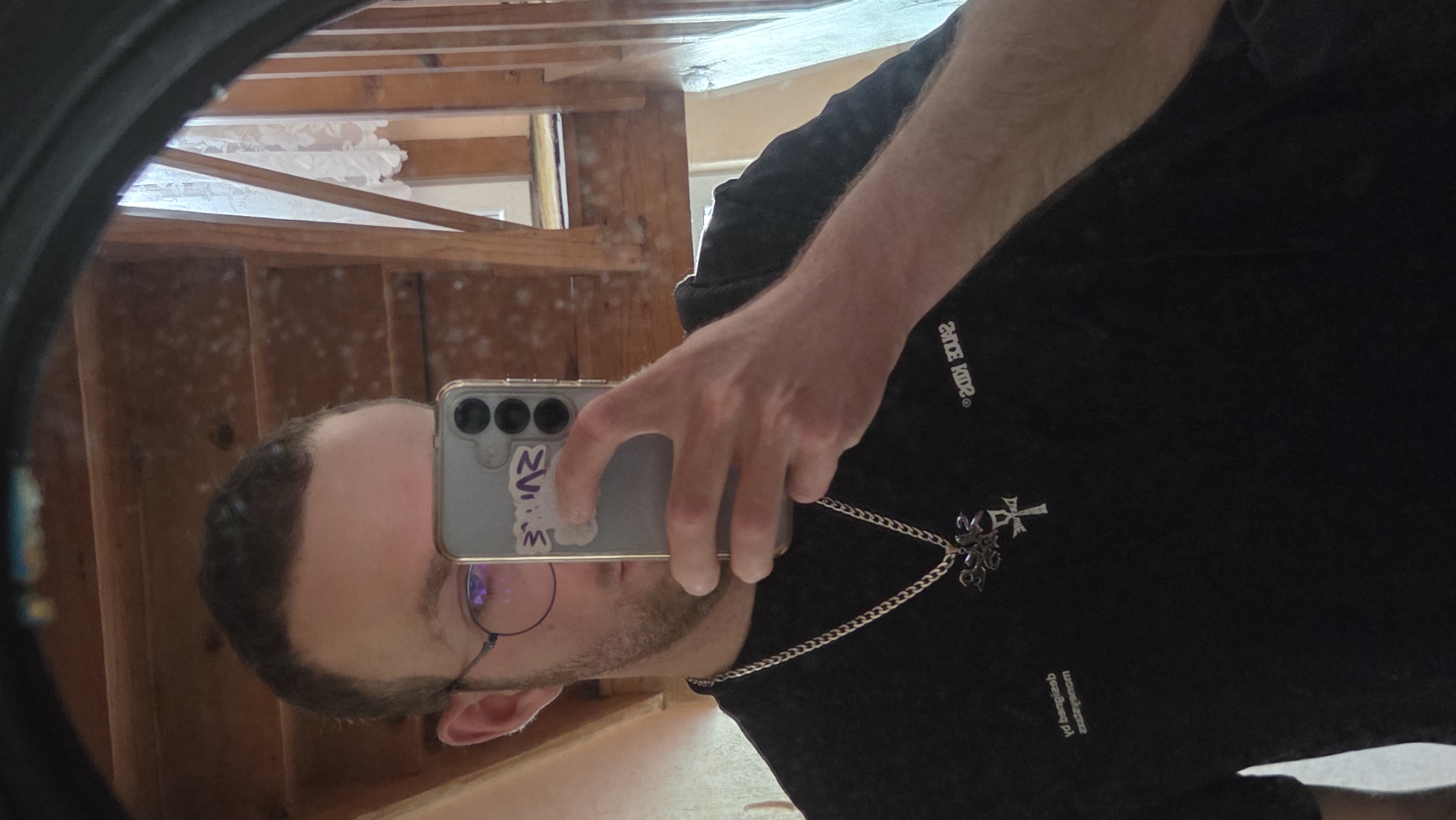 A reflected image of a man taking a selfie is captured in an oval mirror. The man has dark hair and is wearing glasses. He is holding a smartphone, and the phone case has a white sticker on it. He is wearing a dark-colored hoodie, adorned with a silver necklace featuring a decorative pendant. The reflection reveals a room with unfinished wood paneling and a white decorative piece near the ceiling. The specific location in the city of Unknown, Unknown, is not discernible.