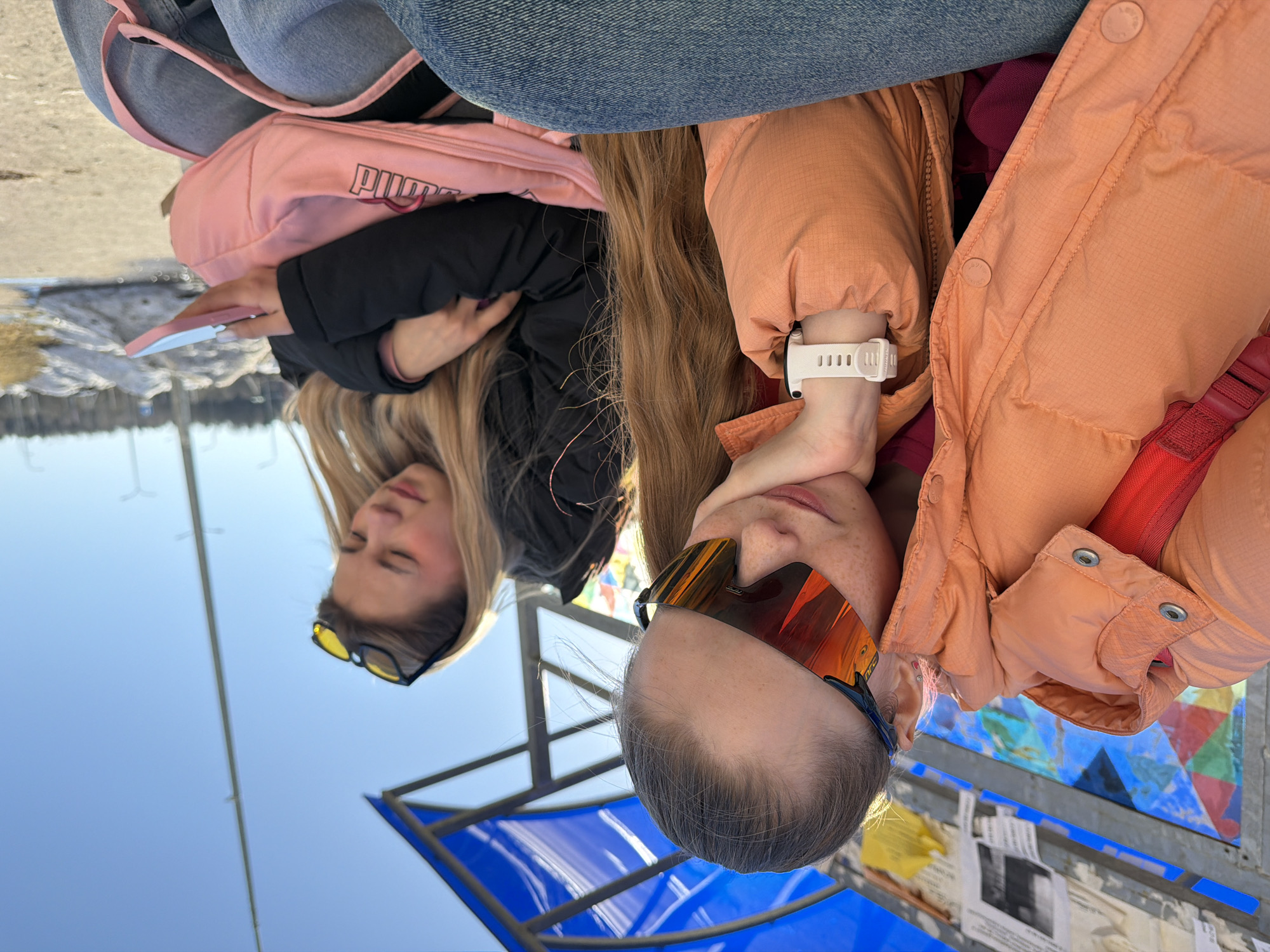 Two women sit outside in Berezniki, Russia. One woman wears orange mirrored sunglasses, an orange jacket, and a white watch. The other woman wears black sunglasses, a black jacket, and blue jeans and holds a pink phone. A pink Puma bag rests near her. A blue metal structure is visible, along with other structures and documents.