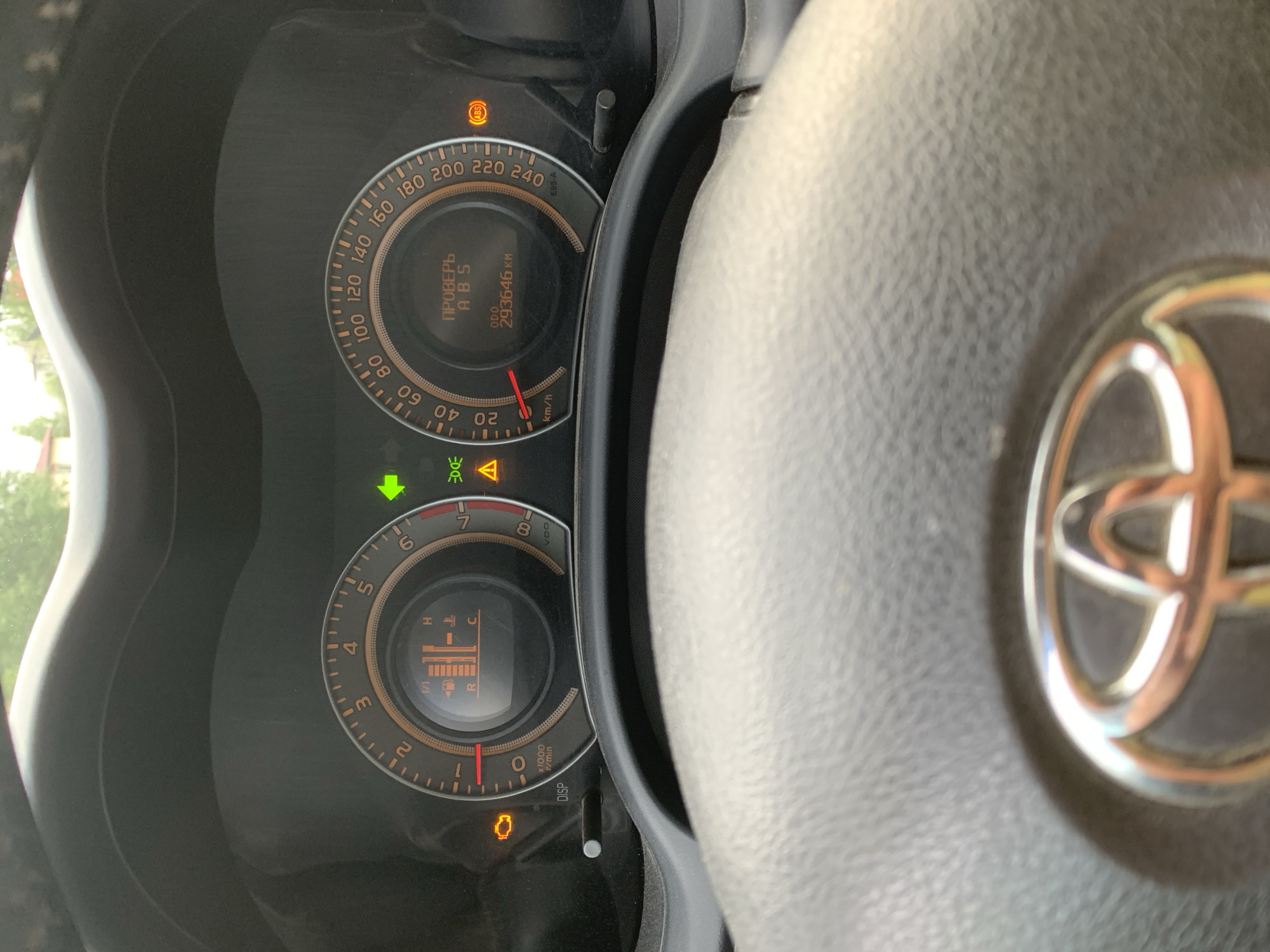 This is a close-up, vertically oriented photograph of a car's dashboard. The primary subjects are the speedometer, tachometer, and a digital display screen, all integrated into the driver's instrument cluster. A portion of the steering wheel, featuring the Toyota logo, is visible on the right side of the frame.
The speedometer is on the left, with markings up to 240 km/h, and the needle points to approximately 20 km/h. To the right of the speedometer is the tachometer, with markings up to 8 x 1000 rpm, and its needle is near the bottom, indicating low engine RPM. In the center of the instrument cluster is a digital display screen showing the word "ПРОВЕРЬ" (which translates to "CHECK" in Russian) and "ABS" in Cyrillic, along with "ODO" and a mileage reading of "293646 км" (293646 kilometers).
Several warning lights are visible: an orange ABS light is illuminated at the top, a green left turn signal is lit, a yellow hazard/warning light is on, and a yellow engine warning light is illuminated near the bottom. The digital display between the tachometer and the speedometer shows gear selection, with indicators for "H" (likely Neutral), and "R" (Reverse), as well as a fuel gauge.
The overall scene is the interior of a car, presumably during daylight hours given the visible reflections and ambient light. The focus is on the dashboard, suggesting a potential issue with the car's ABS system or an overall system check. There are no people visible in the frame. The setting is a car interior.