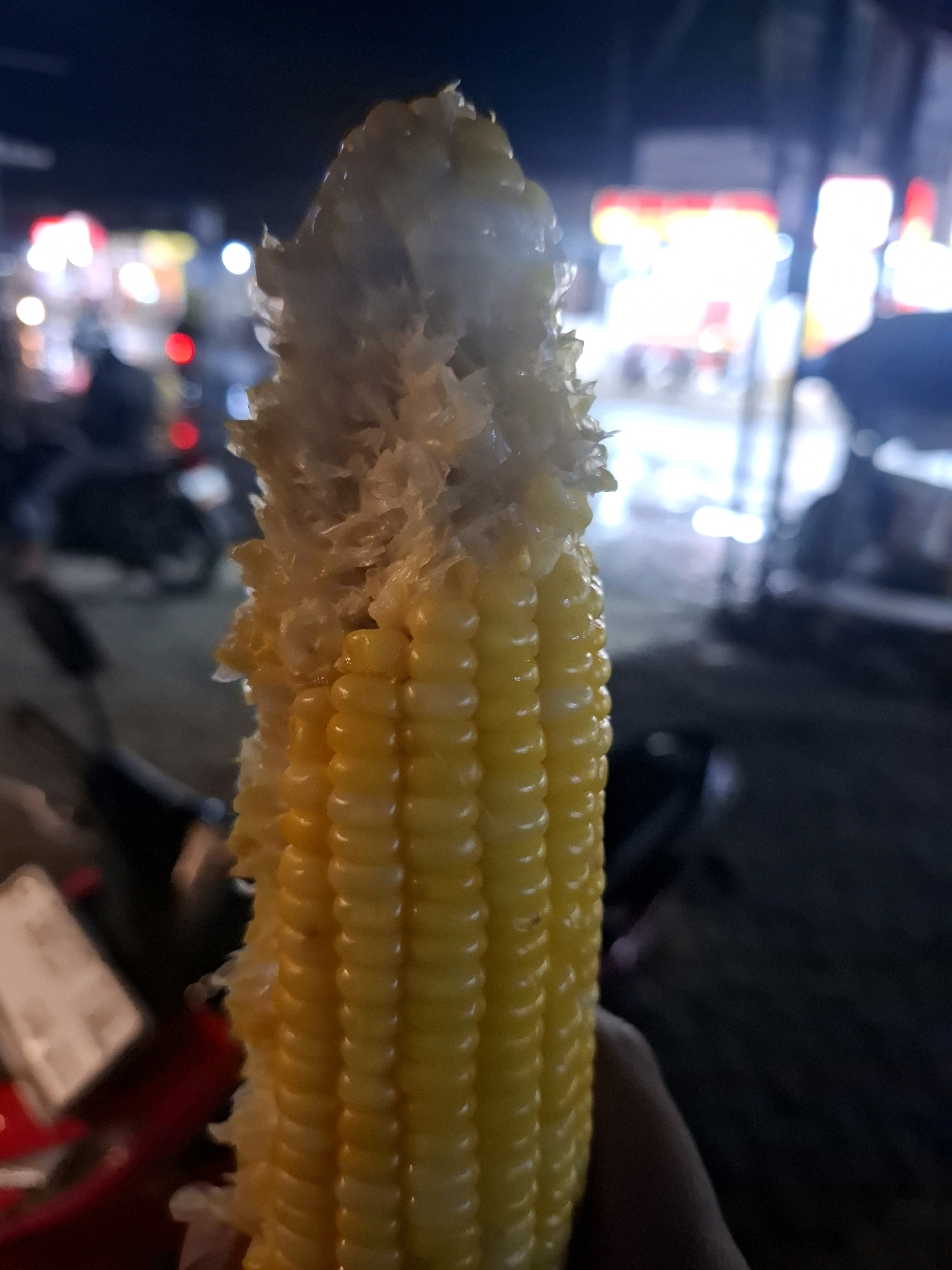 The image shows a close-up of a partially eaten ear of corn held by a person's hand, with a blurred background suggesting an outdoor setting at night. The corn is bright yellow with visible kernels, and the top part appears to be chewed or broken. In the background, out-of-focus lights and the silhouettes of what appear to be motorcycles and possibly street stalls indicate a public area, likely a market or street in the city of Ciheulang Tonggoh, Indonesia. The overall impression is of a casual, urban night scene.