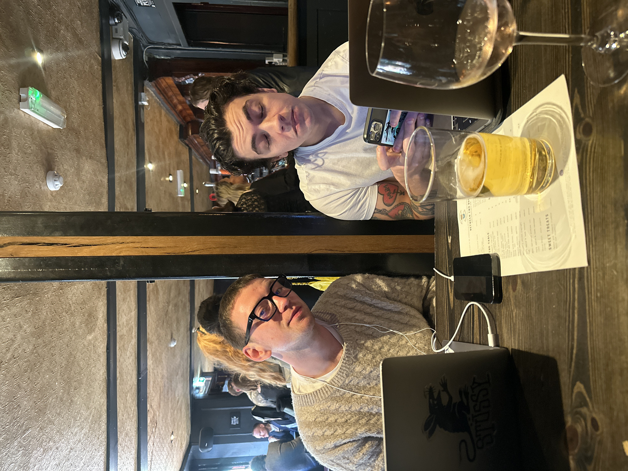 The image depicts two men sitting at a wooden table inside a pub or bar. One man, wearing a white t-shirt and sporting tattoos on his arm, is looking at his mobile phone. The other man, wearing glasses and a light-colored knit sweater, is looking forward. Both have laptops in front of them. On the table, there are two glasses: one containing a yellowish liquid and another a clear liquid. A menu and a charging block are also visible on the table. Background elements include pub patrons and wall fixtures.