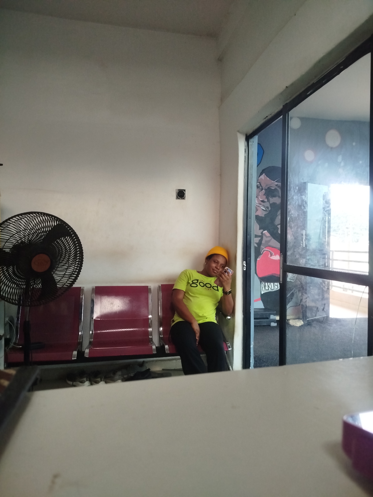 In Uselu, Nigeria, a young woman wearing a neon green t-shirt with the words "the GOOD life" printed on it, and a yellow hard hat, is seated in a waiting area. She is looking at her phone, which she holds in her right hand, and appears to be leaning against the wall. In front of her are three burgundy waiting chairs with metal frames, and to her left, a black oscillating fan is positioned. Underneath the chairs, a pair of dark shoes and what appear to be sandals are placed on the floor. The waiting area has white walls, and a large glass window to the right offers a view of an interior space with a mural of a boxer. The overall environment suggests a casual or waiting space, possibly within a facility or office building.