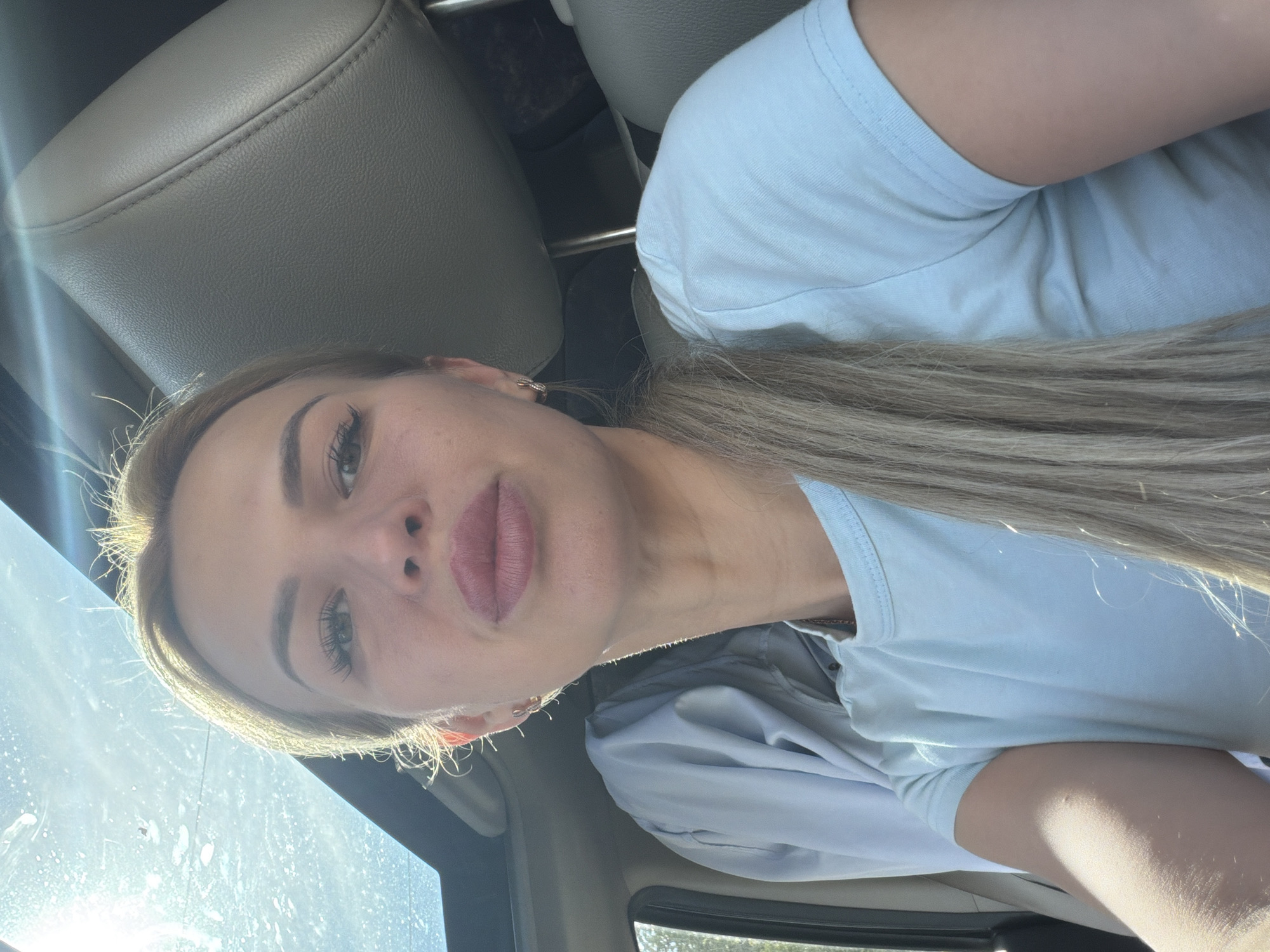 A blonde woman is pictured from the chest up, inside a car. She is looking directly at the camera with her lips slightly parted. She has on a light blue t-shirt, and her hair is pulled back in a ponytail. The sun is shining through the sunroof of the car, creating a bright glare. The woman appears to be in good spirits. The car appears to be parked in a residential area in Kyiv, Ukraine.