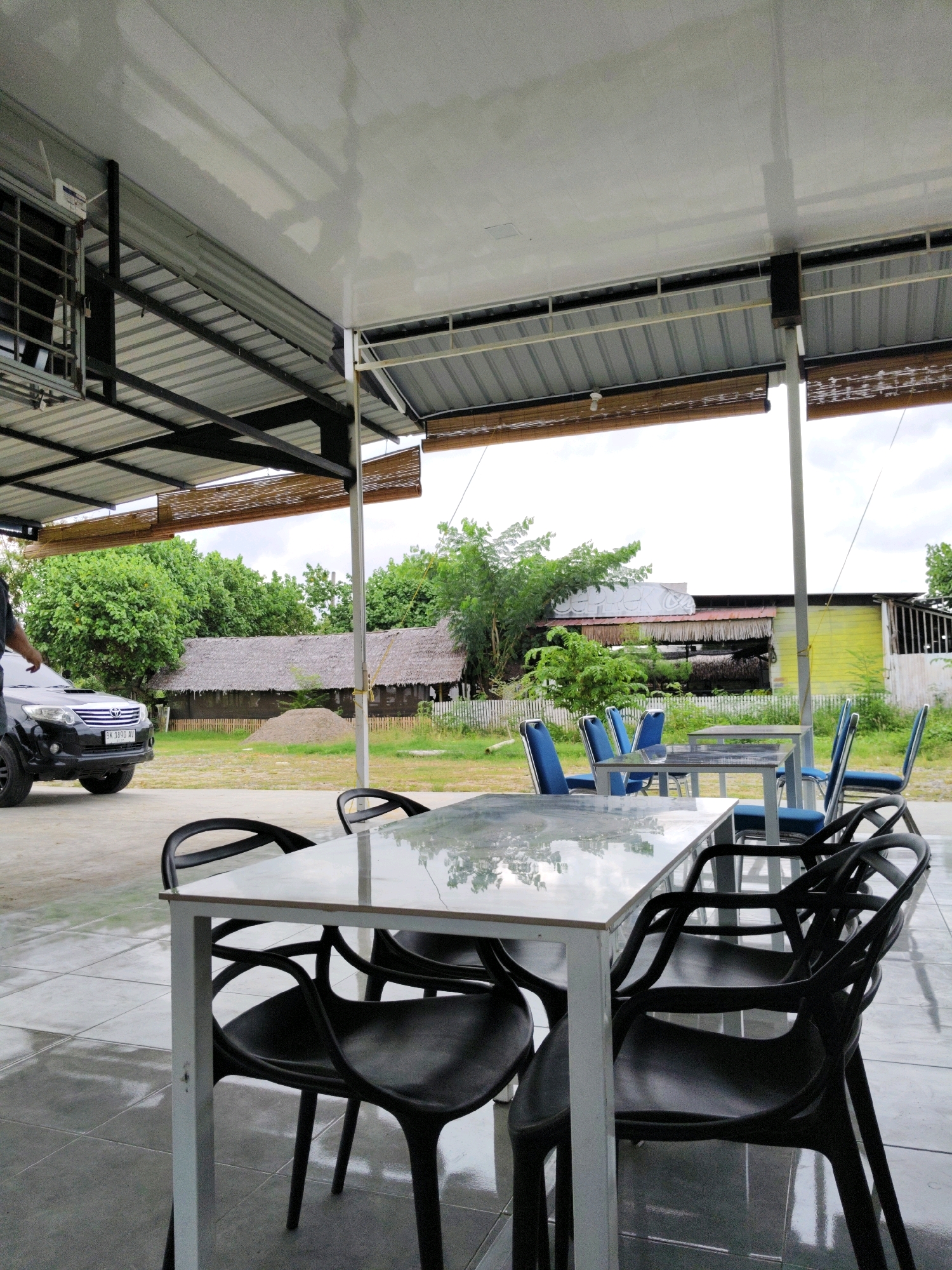 The image depicts an outdoor seating area, likely a cafe or restaurant, in Lhokseumawe, Indonesia. The foreground is dominated by a reflective table with black chairs, and a second table with blue chairs is visible further back. Beyond the seating area, a black SUV is parked. In the background, traditional Indonesian houses with thatched roofs are set against a backdrop of lush green trees. The overall scene suggests a relaxed, tropical atmosphere, possibly in a rural or semi-rural setting within the city. The sky is overcast, indicating a possibly humid or rainy day.