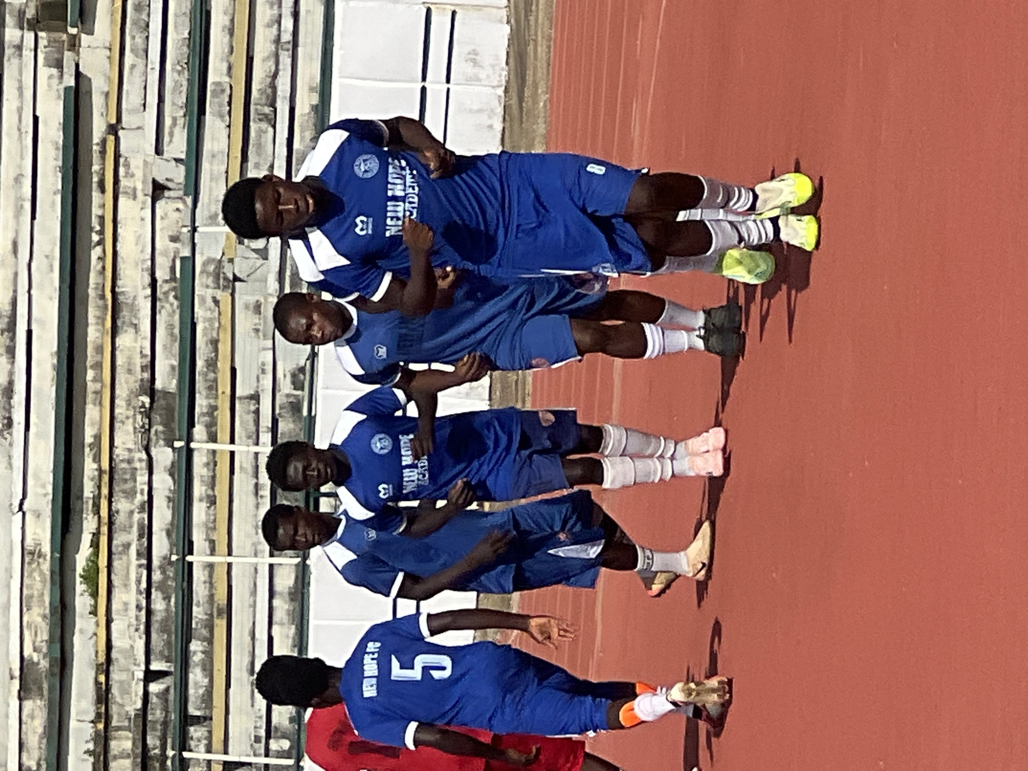 In the city of New Stadium, Nigeria, five young male soccer players are captured on a red running track with a stadium seating area in the background. The players are wearing matching blue and white uniforms, with one player distinguished by a red jersey. The player on the far left is wearing a blue jersey with the number "5" on the back, and the text "NEW HOPE FC". He is also wearing orange socks and cleats. The player next to him is wearing a blue jersey with "NEW HOPE ACADEMY" written on it. The player in the center is also in a blue uniform. The player to his right is wearing a blue jersey with the number "8" on the back. The player on the far right is in a blue uniform. All players appear to be in motion, suggesting they are either warming up or engaged in a training exercise. The stadium seating is visible in the background, appearing weathered and partially occupied by shadows. The lighting indicates it is daytime, likely a sunny day, given the sharp shadows cast by the players.