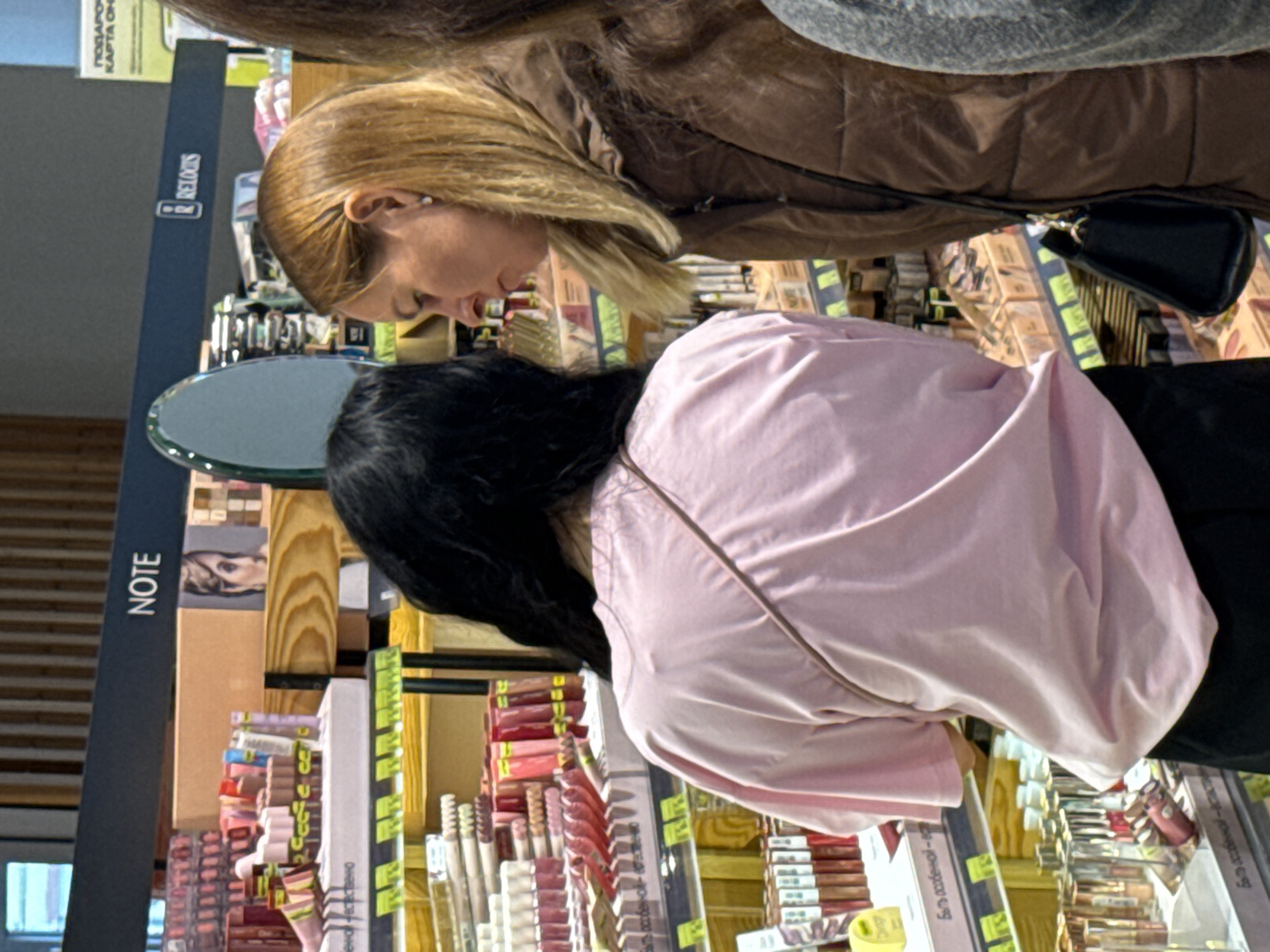 The image captures two women browsing in what appears to be a cosmetics or beauty section of a retail store. The scene is indoors, likely during daytime operating hours.
In the foreground, with her back mostly to the viewer, is a woman with long dark hair, wearing a light pink short-sleeved top and dark-colored bottoms. She is bent over, attentively looking at products displayed on a lower shelf.
Behind her, slightly to the right, is another woman with long blonde hair, wearing a brown quilted jacket. Her profile is visible as she also looks down at the products on the shelves. A small black rectangular purse with a gold zipper is slung over her shoulder.
The store environment features multiple display shelves packed with various cosmetic items, predominantly lip products and other makeup, arranged neatly in rows. Many products have small, yellow-ish labels, likely price tags or product information. A small, oval-shaped mirror with a metallic frame is visible on one of the display stands between the two women.
Several brand names and texts are visible on vertical signage. On a dark blue sign, the brand "RELOUIS" is clearly legible. Further down and to the left, another dark blue sign displays the brand "NOTE". In the upper left corner, partially visible, are Russian words "ПОДАРОЧНАЯ КАРТА ОН...", which translates to "GIFT CARD ON...". Other Cyrillic text can be seen on product labels and price tags, further confirming the location context of Kazan, Russia. The women are engaged in the activity of shopping for beauty products.