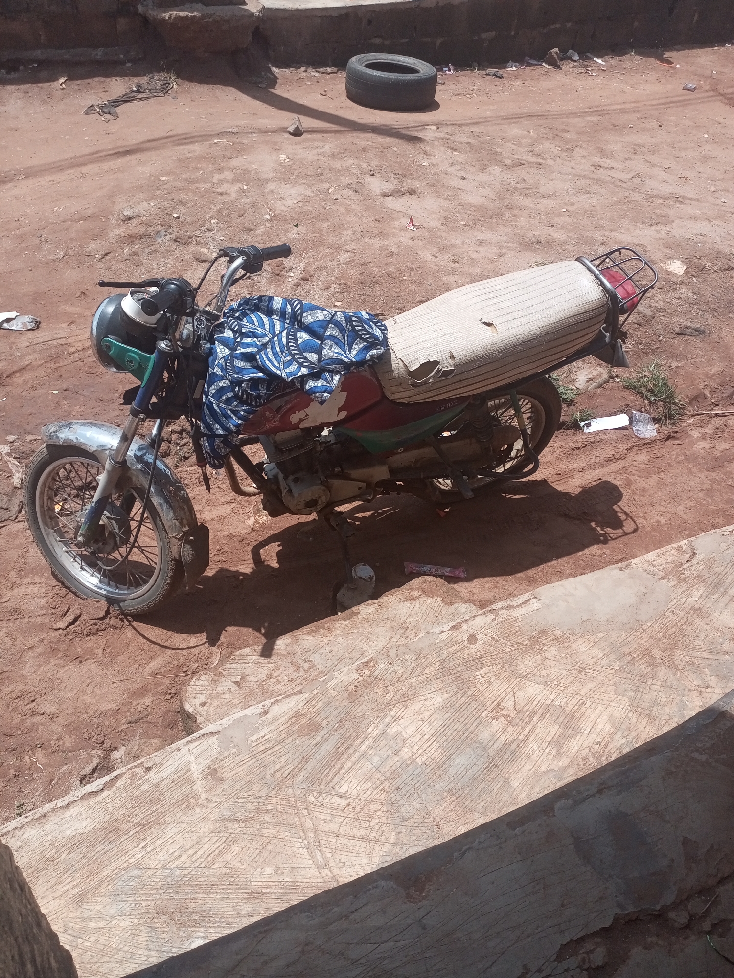 The image captures a parked, well-used utility-style motorcycle on an unpaved, reddish-brown dirt surface. Bright sunlight illuminates the scene, casting distinct and relatively short shadows, suggesting it is midday or early afternoon on a clear, dry day.
The motorcycle is the central subject. A blue and white patterned cloth is draped over its handlebars and the front section of the seat. The long, light-colored (beige or off-white) motorcycle seat shows considerable wear and tear, with several visible rips. The front fender is chrome, appearing scuffed and dirty. Near the engine on the side of the motorcycle's frame, a small label or sticker is partially visible with the text "BM 100".
The ground is uneven, composed of loose dirt, small stones, and some sparse dry vegetation. In the background, there's a raised, somewhat rugged structure of dirt and concrete, with an old black tire resting on the ground. The foreground features a wide concrete step or ledge, heavily marked with numerous crisscrossing scratch lines.
No people are visible in the image, and the motorcycle appears to be stationary, indicating no active event or activity. The overall setting suggests an outdoor, possibly semi-urban or rural environment, characterized by unpaved areas and worn structures.