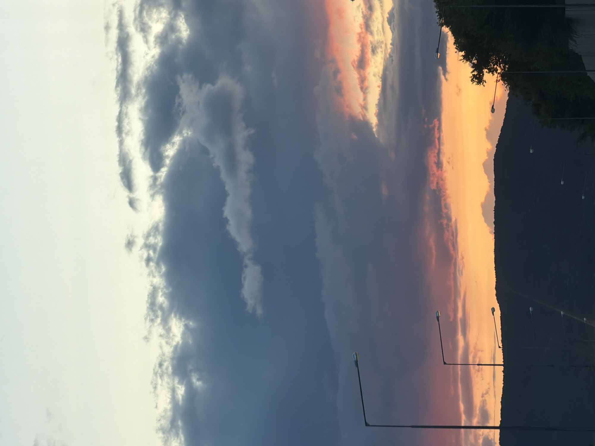 The image depicts a twilight scene with a dramatic sky. Dark, heavy clouds dominate the upper portion of the frame, gradually giving way to a band of vibrant orange and pink hues near the horizon, illuminated by the setting sun. Silhouetted against this colorful sky are the dark outlines of rolling hills. Several streetlights, tall and slender, are visible in the foreground and midground, their lamps glowing faintly. To the right, a dense line of dark green trees stands out against the sky, suggesting a treeline or a wooded area. The overall impression is one of a natural landscape at dusk, with man-made elements like streetlights interspersed. There are no people or animals visible in the image. The scene appears to be an outdoor location, possibly a highway or a road leading into or out of a suburban or natural area. There is no specific indication of the city of Samara, Russia within the visual content of the image.