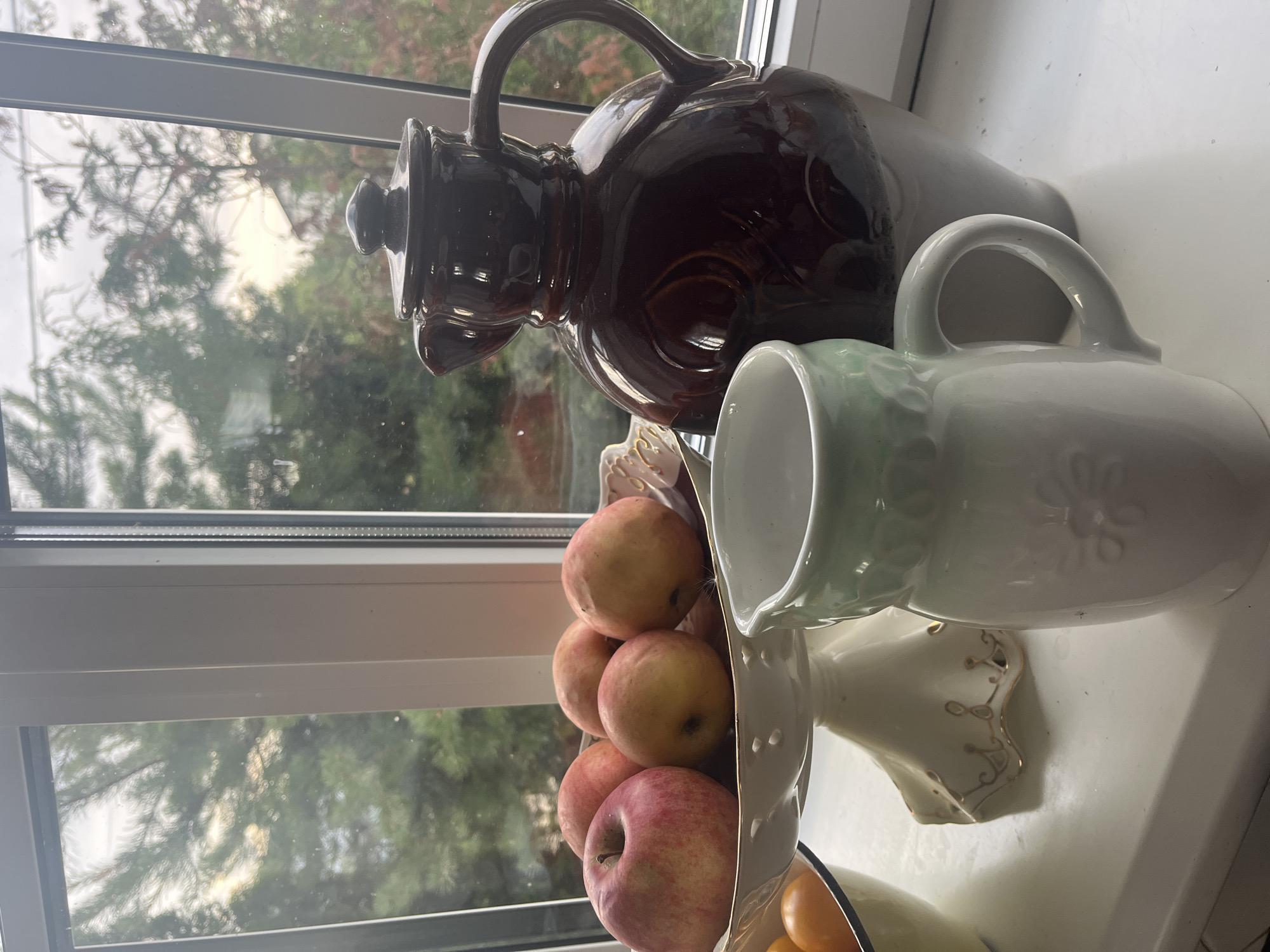 The image shows a still life arrangement on a windowsill overlooking a natural scene. In the foreground, a white ceramic pitcher with a pale green rim sits in front of a large, dark brown ceramic jug. To the left, a white pedestal bowl holds several red and yellow apples. Partially visible to the far left is a yellow bowl containing oranges. The windowsill is white and reflective. The background, visible through the window, shows trees with a mix of green and autumnal foliage, with a hint of a building or structure in the distance. The lighting suggests it is daytime, possibly late afternoon or early morning, with a diffused light coming from the sky. The scene is indoors, likely in a domestic setting in Vladimirovka, Russia.
