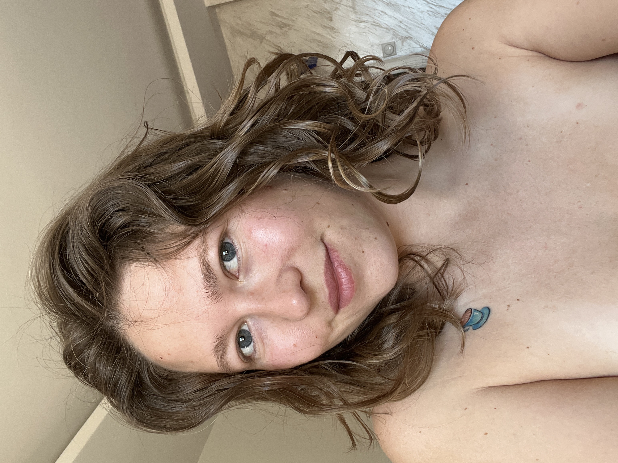 A head and shoulders shot of a light-skinned person with wavy, shoulder-length brown hair and blue eyes. A small blue tattoo of a coffee cup is visible on the upper chest area. The background appears to be a wall with a light fixture, and a textured surface that might be tile. No information is available that indicates this image was taken in Podgorica, Montenegro.