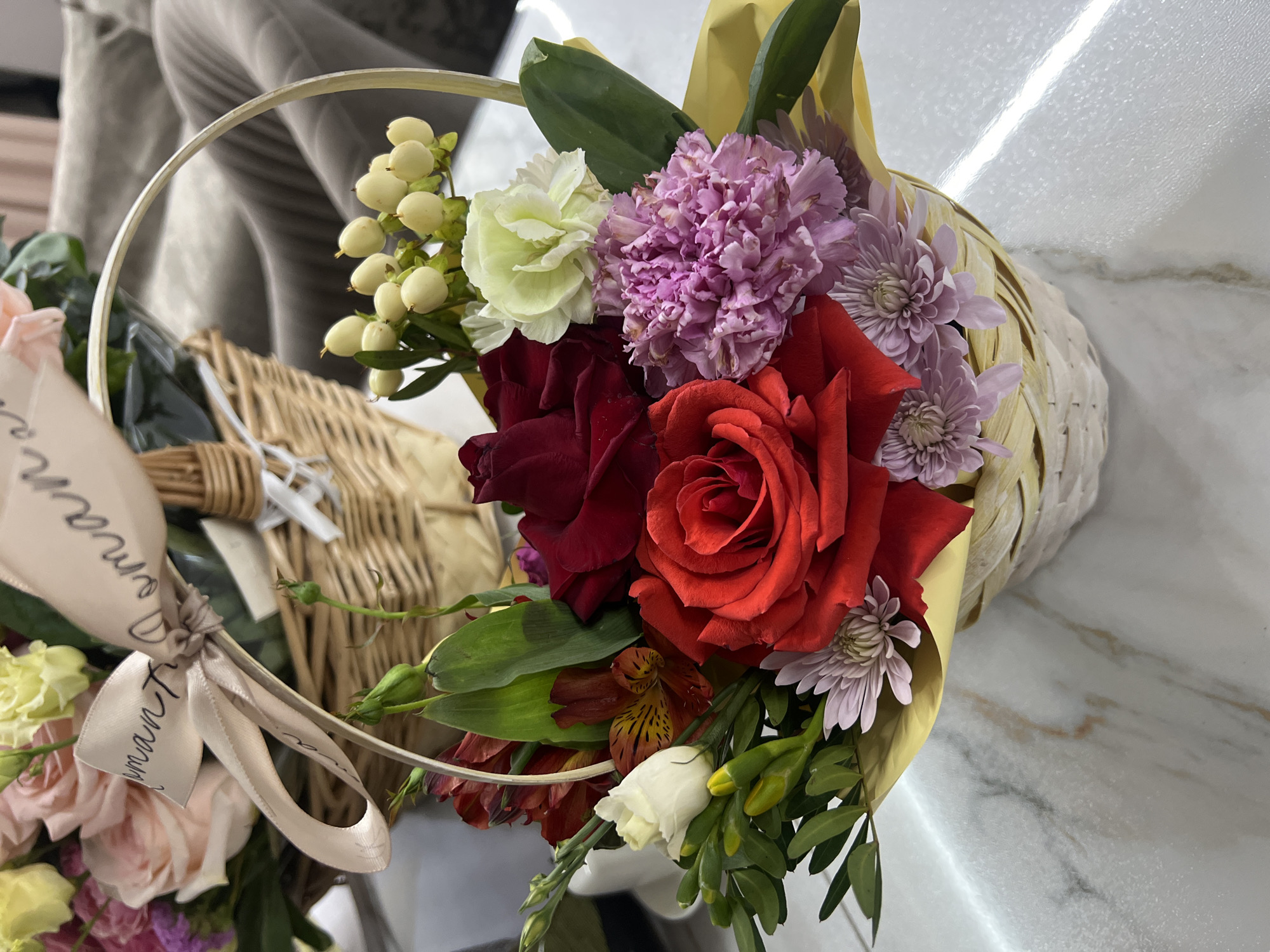 A floral arrangement in a basket with a handle sits on a marble-patterned surface. The arrangement includes red roses, purple carnations, white berries, white and burgundy alstroemeria, and other flowers. The basket is wrapped in yellow paper. A ribbon with writing on it is tied around the handle of the basket. The setting appears to be indoors. It cannot be confirmed that the location is in Tyumen, Russia.