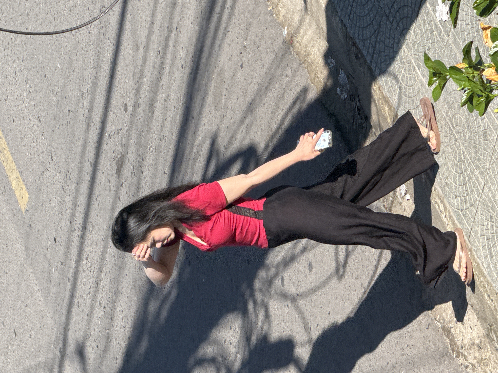 A woman with long, dark hair, wearing a red shirt with black accents and black pants, walks on a paved city street in Da Lat, Vietnam. She is holding a smartphone in her right hand and shielding her eyes with her left hand, suggesting she is looking up or into the sun. The street is gray asphalt with a yellow line painted on it. Shadows from trees or buildings create a dappled pattern on the road. A sidewalk with a geometric pattern is visible to the right of the street. In the foreground, on the sidewalk, there are green plants and some scattered debris. The overall lighting suggests it is a sunny day.