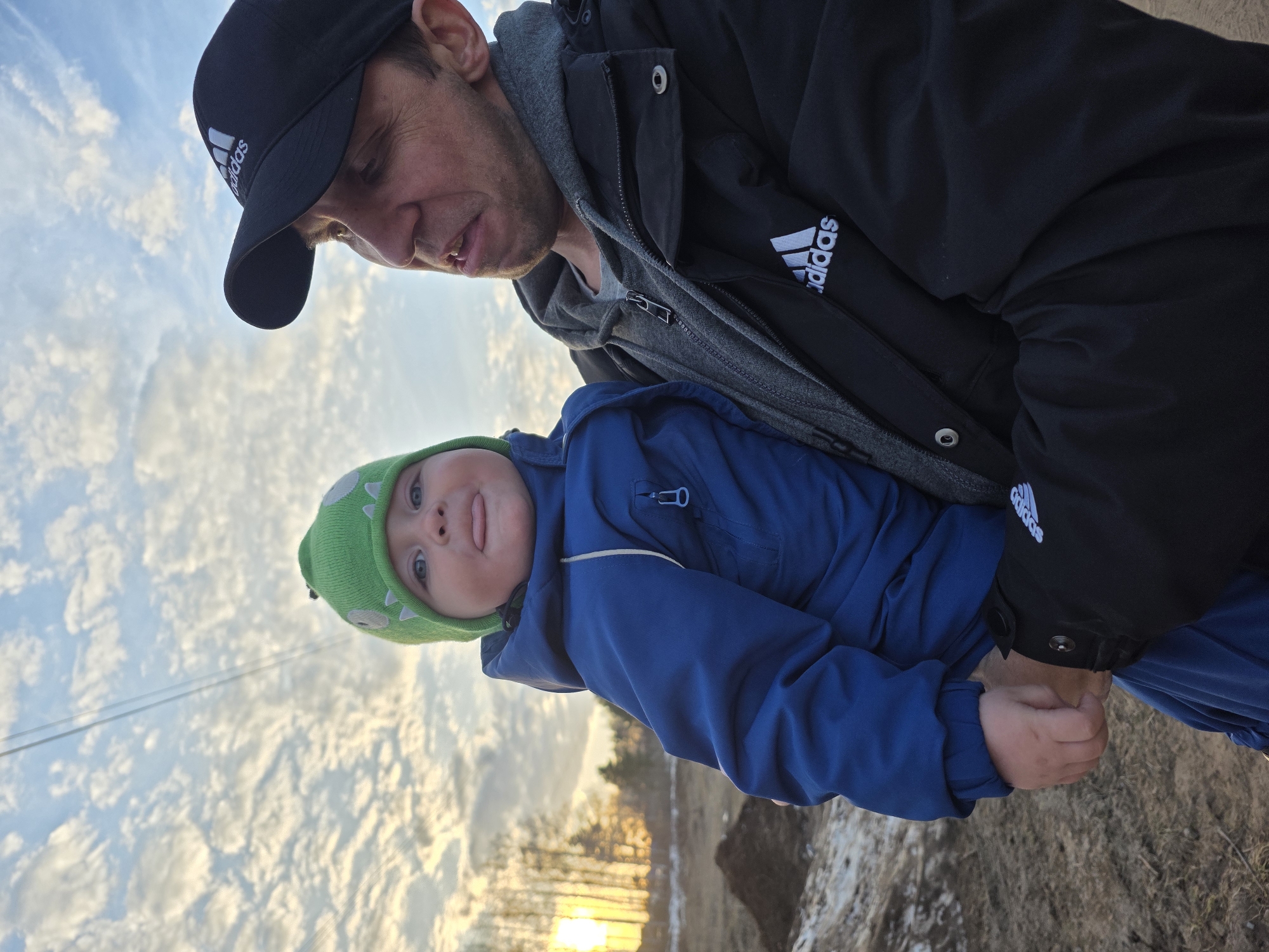 A man wearing a black Adidas baseball cap and a black Adidas jacket with a gray hoodie underneath holds a baby in a blue jacket and a green dinosaur-themed hat. The baby is sticking out their tongue. The setting is outdoors with a backdrop of a cloudy sky and the beginning of a sunset over trees. The ground appears to be somewhat muddy and partially covered in snow or frost. The exact location within Noviy, Russia is not discernable from the image.