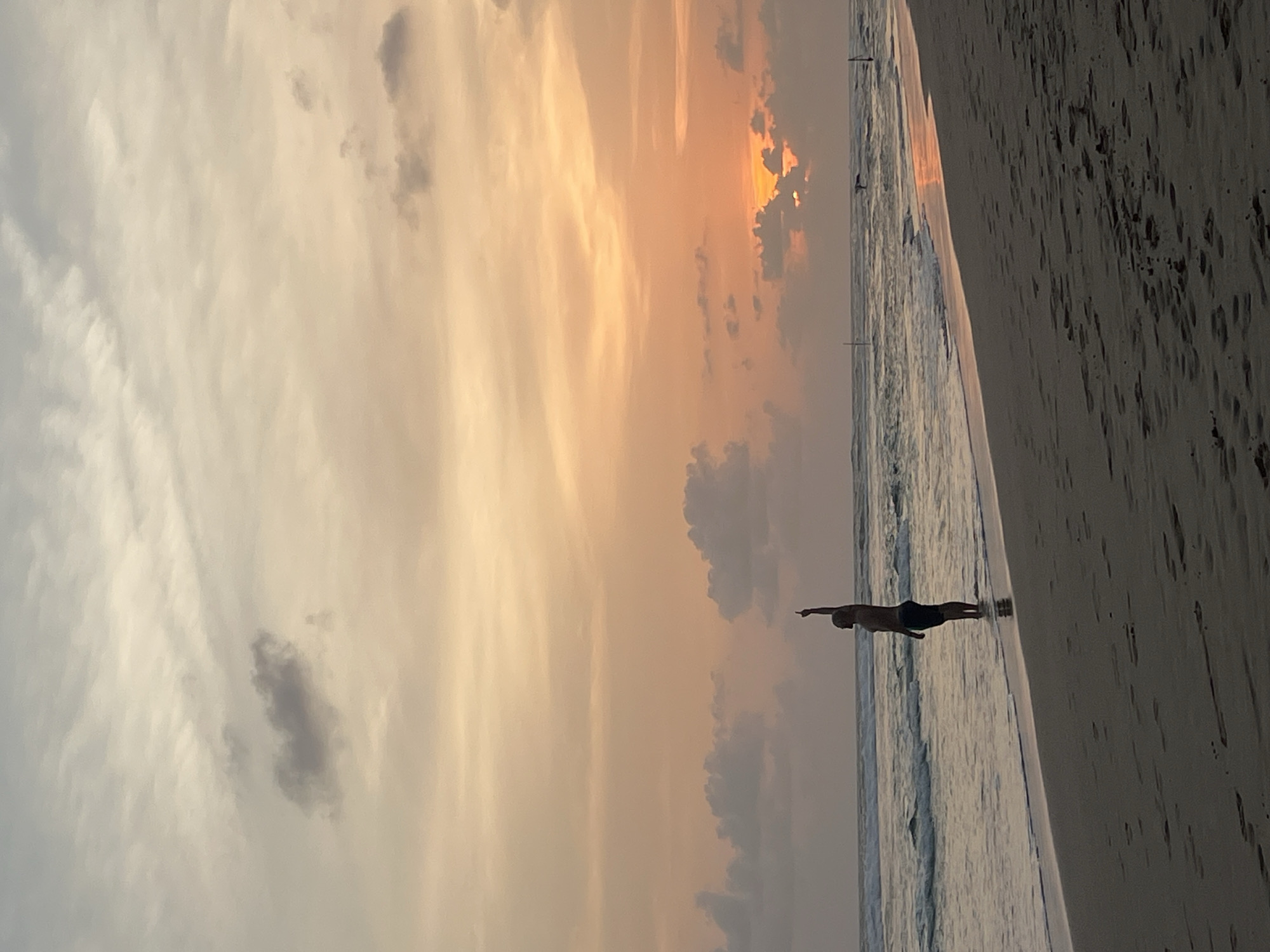 A horizontal, outdoor shot captures a person standing near the edge of a beach in Kathaluwa, Sri Lanka, at dusk or dawn. The sky is a mix of grey and pale orange clouds, suggesting a sunrise or sunset. A body of water runs parallel to the beach, with small waves visible near the shore. The sand appears dark and textured. The person is silhouetted and is facing the ocean with one arm outstretched.