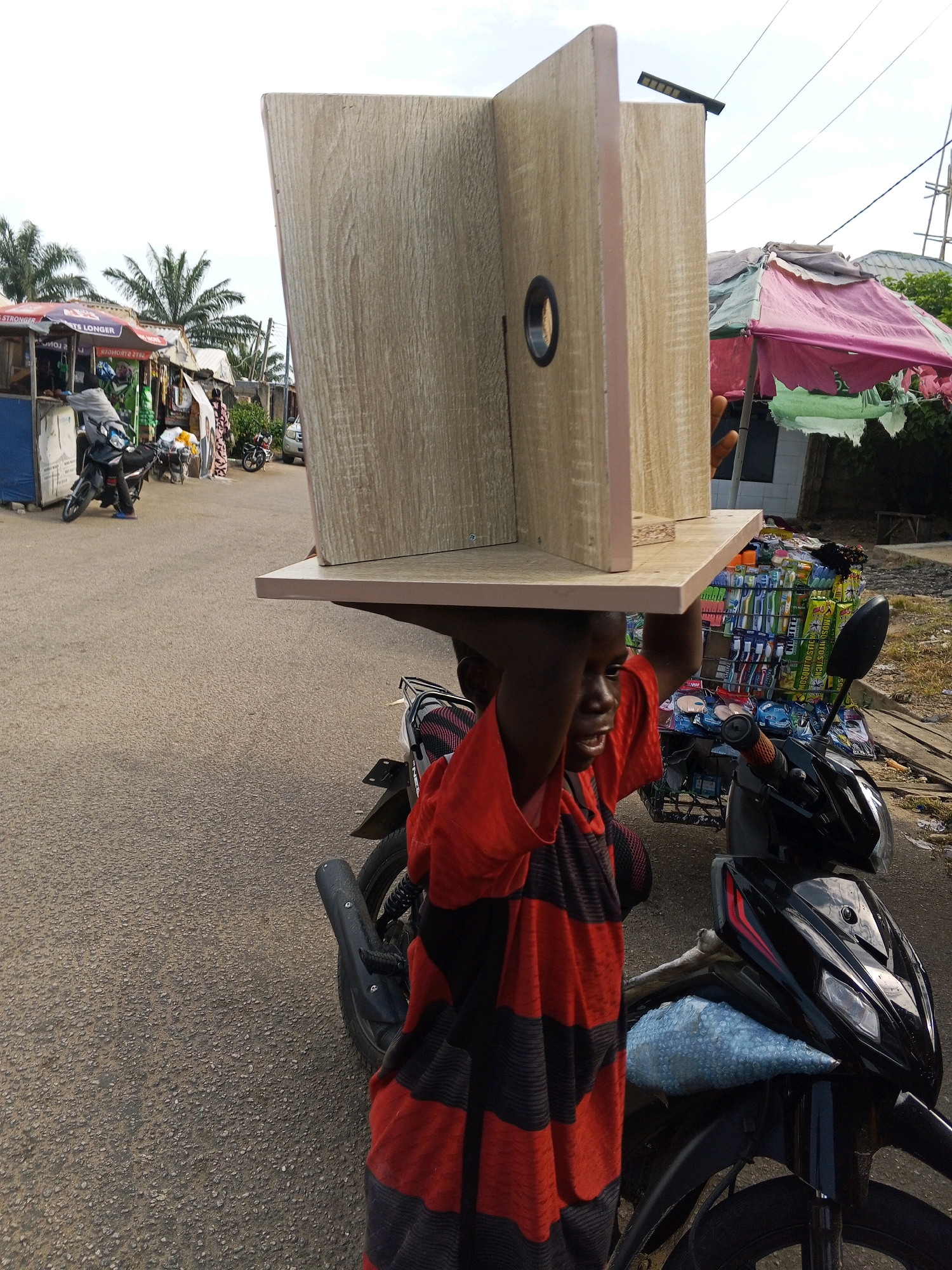 In Lokoja, Nigeria, a young boy carries a large, wooden, cabinet-like structure on his head. He is wearing a red and black striped t-shirt and is walking on a paved road. To his left, there is a market stall with various goods for sale, including toothbrushes. A black motorcycle is parked next to him, and a blue bubble-wrapped object is visible on its front. In the background, there are more market stalls, palm trees, and buildings, suggesting a busy urban environment.
