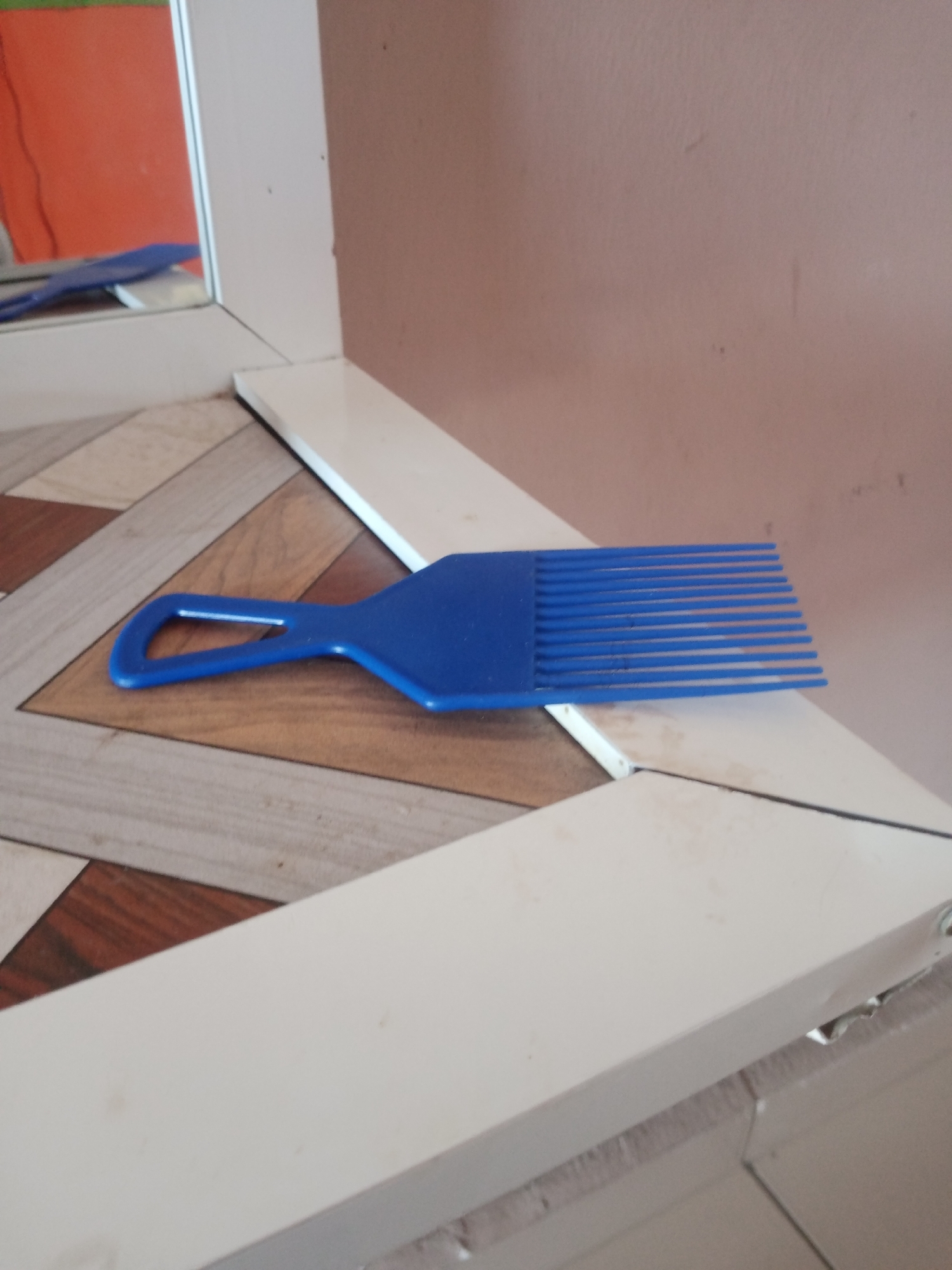 A blue plastic afro pick is lying horizontally on a white surface, its teeth pointing to the right. The pick has a rectangular handle with a looped opening. The background shows a wall with a pinkish hue and a reflection of an orange surface and a blue object in a mirror. The floor beneath the pick is tiled with a pattern of light brown, beige, and grey diamond shapes. The overall scene appears to be an indoor environment, possibly a home or a salon, located in Jalingo, Nigeria. No people are visible in the image.