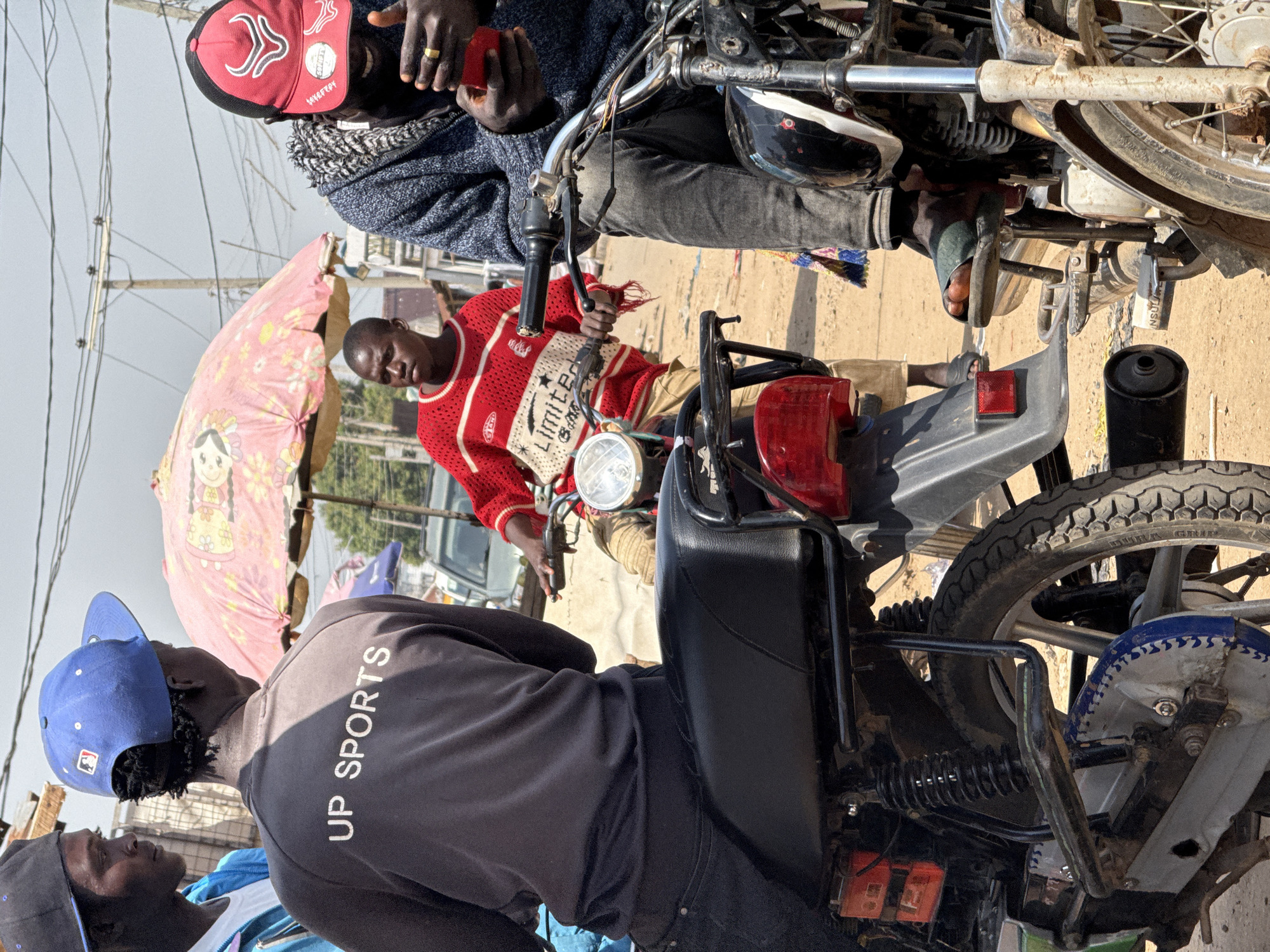 In Abuja, Nigeria, on a dusty street, four young men are gathered around two motorcycles. One man, wearing a blue baseball cap and a gray shirt with "UP SPORTS" printed on the back, is seated on the back of a motorcycle. Beside him, another man in a red and white sweater stands. To the right, a man in a red baseball cap is seated on a motorcycle, looking at his phone. A fourth man, wearing a blue jacket, stands in the background, looking towards the left. An open umbrella with a floral pattern stands behind the men, providing shade. The scene suggests a casual street gathering or waiting area, typical of informal commerce or transportation hubs in the city.