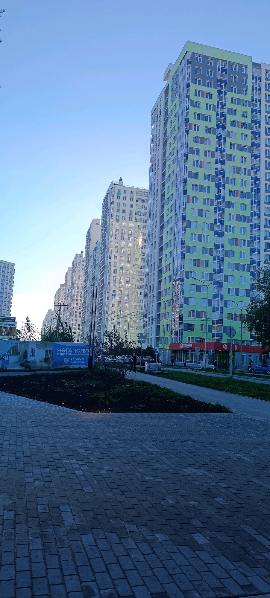 The image depicts a modern residential development in Perm, Russia, specifically the "Megapolis" microdistrict. In the foreground, a paved walkway made of dark grey rectangular pavers leads towards the street. To the left, a landscaped area with dark soil and some sparse vegetation borders the walkway.
Dominating the mid-ground and background are several tall, multi-story apartment buildings. The building on the far right has a striking facade of alternating light green and dark blue panels, with numerous windows. To its left, a row of white apartment buildings stretches into the distance, each featuring a grid of evenly spaced windows, some of which are illuminated, suggesting occupancy. The architectural style is contemporary, characterized by clean lines and large glazed areas.
On the street level, in front of the buildings, a sidewalk runs parallel to the road. Several cars are visible parked along the street and in a parking lot. A blue car is moving away from the viewer on the right side of the street. A red and white storefront with prominent signage is visible at the base of the green and blue building. Adjacent to it, a blue billboard displays text in Cyrillic, including "МЕГАПОЛИС застройщик" (Megapolis Developer) and a phone number.
Two individuals are walking on the sidewalk near the billboard. One appears to be wearing dark clothing, and the other is further in the background. The sky is clear and light blue, indicative of daytime, possibly late afternoon or early morning due to the soft lighting. The overall impression is that of a new, developing urban residential area.
