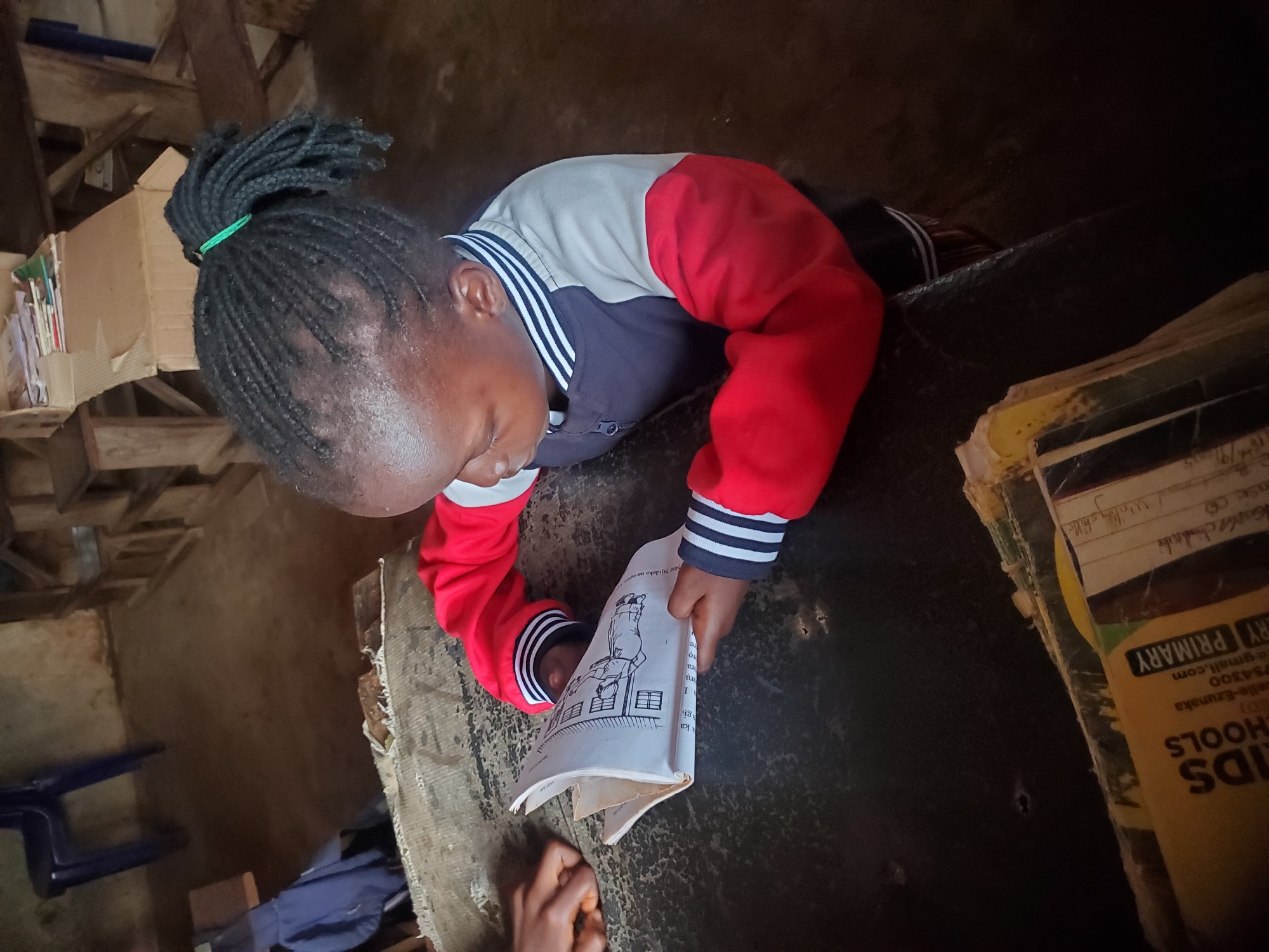 A child with braided hair tied with a green band is shown reading a book, presumed to be a student at a school in Ukwu-Abwa, Nigeria. The child is wearing a jacket with red and gray sleeves with black and white striped cuffs and collar. The open book features a line drawing. Additional books and a cardboard box are visible in the background. The environment appears to be a modest interior space, potentially a classroom. A partial view of another person's hand is at the bottom of the image.