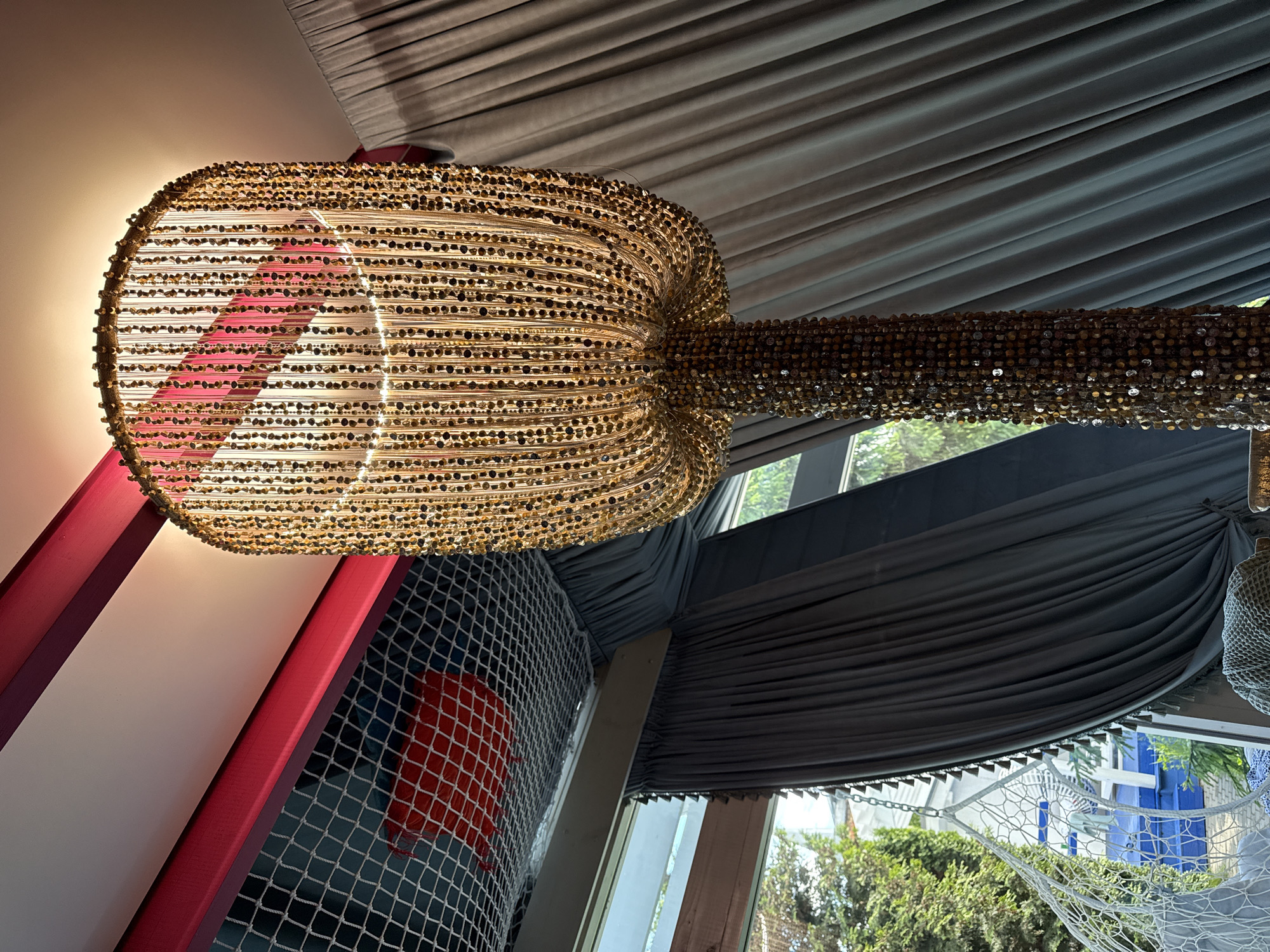 The image shows a decorative light fixture hanging from the ceiling of a room. The fixture is composed of numerous small, reflective pieces, forming a bulbous shape with a long, narrower extension. The fixture is lit from within, casting a warm glow.
The background reveals a wall with red and white stripes, alongside a net-like structure with a red square visible behind it. Beyond this, there is a window showing foliage and architectural elements outside. The ceiling is draped with a dark, pleated fabric.