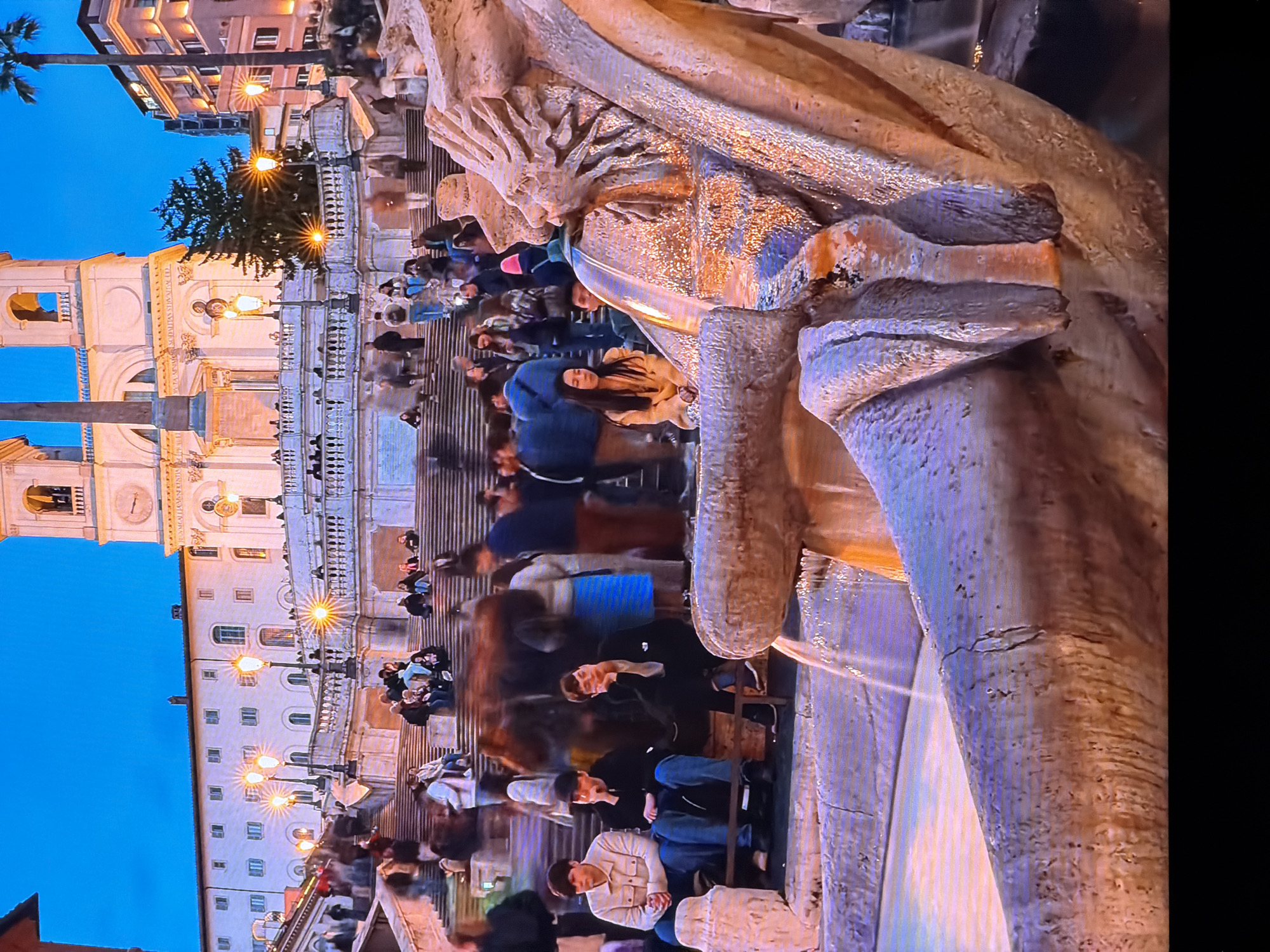 The image depicts a twilight scene at the Spanish Steps in Rome, Italy. In the foreground, a section of the Barcaccia Fountain is visible, with water flowing. Several individuals are seated on the steps, some engaged in conversation or observing their surroundings. In the background, the church of Trinità dei Monti stands prominently at the top of the steps, illuminated by lights. The overall atmosphere suggests a public gathering place, with people enjoying the evening in a historic urban setting. The image is not located in Chirchik, Uzbekistan.