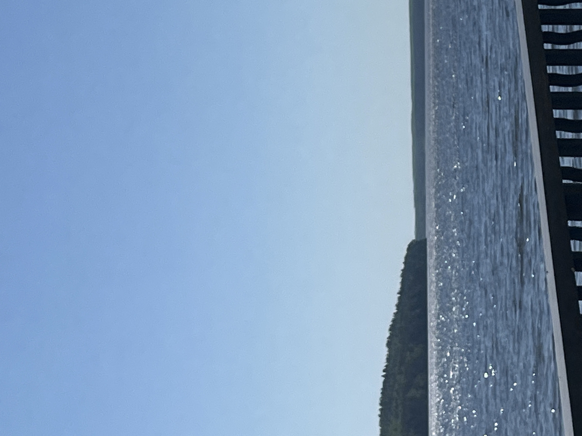 The image shows a portion of a body of water, possibly a lake or reservoir, under a clear blue sky. Vegetation or a forest line is visible along the water's edge. A structure, possibly a pier or boardwalk, with wooden slats is visible on the right side of the frame. The location is possibly Chaikovsky, Russia.