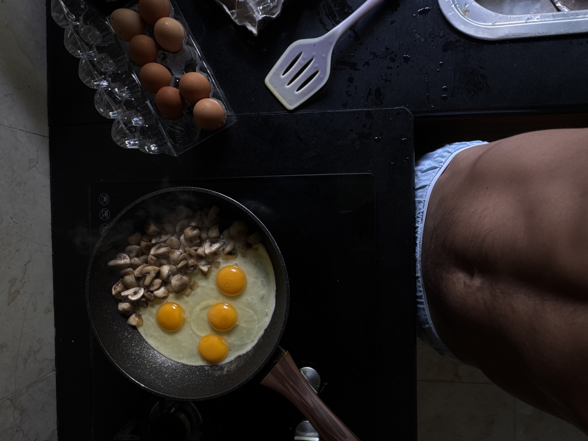 A high-angle, full shot captures a person in light blue underwear preparing eggs and mushrooms for breakfast in a kitchen located in Kuta Selatan, Indonesia. The scene is illuminated by a top-down light source, casting shadows that create a dramatic contrast between light and dark. In the foreground, the lower back and buttocks of the person are visible, their skin showing a light tan and visible body hair. They are wearing light blue underwear with a white elastic waistband.
The main subject of the image is a black frying pan on a black induction stovetop. The pan contains four raw eggs with bright orange yolks and some cooked sliced mushrooms. Steam is rising from the pan, indicating it is on heat. To the right of the pan, an egg carton holding several brown eggs is visible. Further to the right, a light purple spatula lies on the wet stovetop. The stovetop itself appears to have water droplets scattered across its surface. In the background, a tiled wall can be seen, and to the far right, a glimpse of a stainless steel sink is visible. The overall impression is one of a candid, intimate moment in a domestic setting.