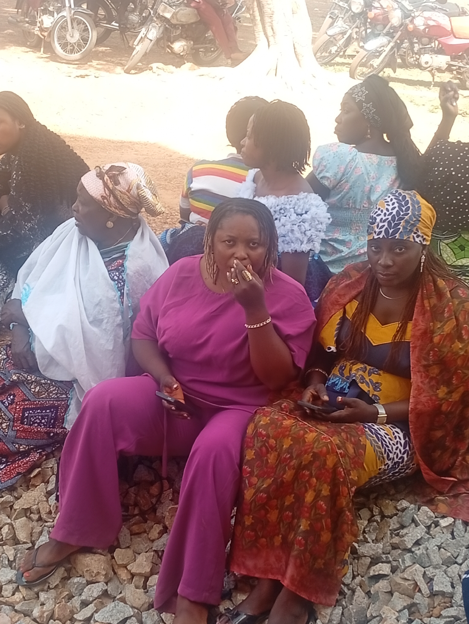 The image depicts a group of women sitting outdoors in Unknown, Unknown. Four women are in the foreground, sitting on what appears to be a rocky or gravelly ground. The woman in the center is wearing a magenta outfit, the woman to her right is wearing a headscarf and a colorful patterned dress with an orange shawl, and the woman to her left has a patterned headscarf and a white shawl. Each are holding a phone. Behind them are other women with various clothing styles and patterns. Motorbikes are visible in the background along with a tree. The environment appears to be an outdoor gathering or event.
