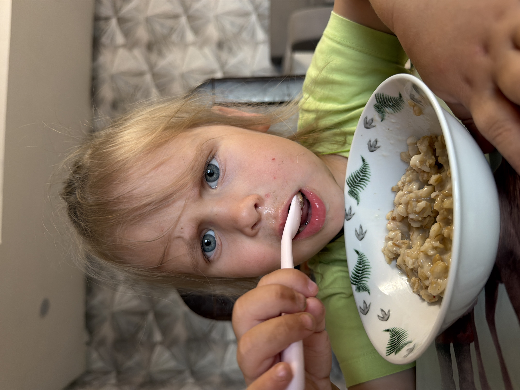 A young girl with light blonde hair and blue eyes is sitting indoors, possibly in a residential setting, as indicated by the patterned wall covering visible in the background. She is holding a pink toothbrush in her mouth, cleaning her teeth, and looking directly at the camera. Next to her is a white bowl decorated with green fern and paw print designs, containing what appears to be a portion of oatmeal. She is wearing a lime green shirt. There is no information to confirm that the photo was taken in the city of Chaikovsky, Russia.