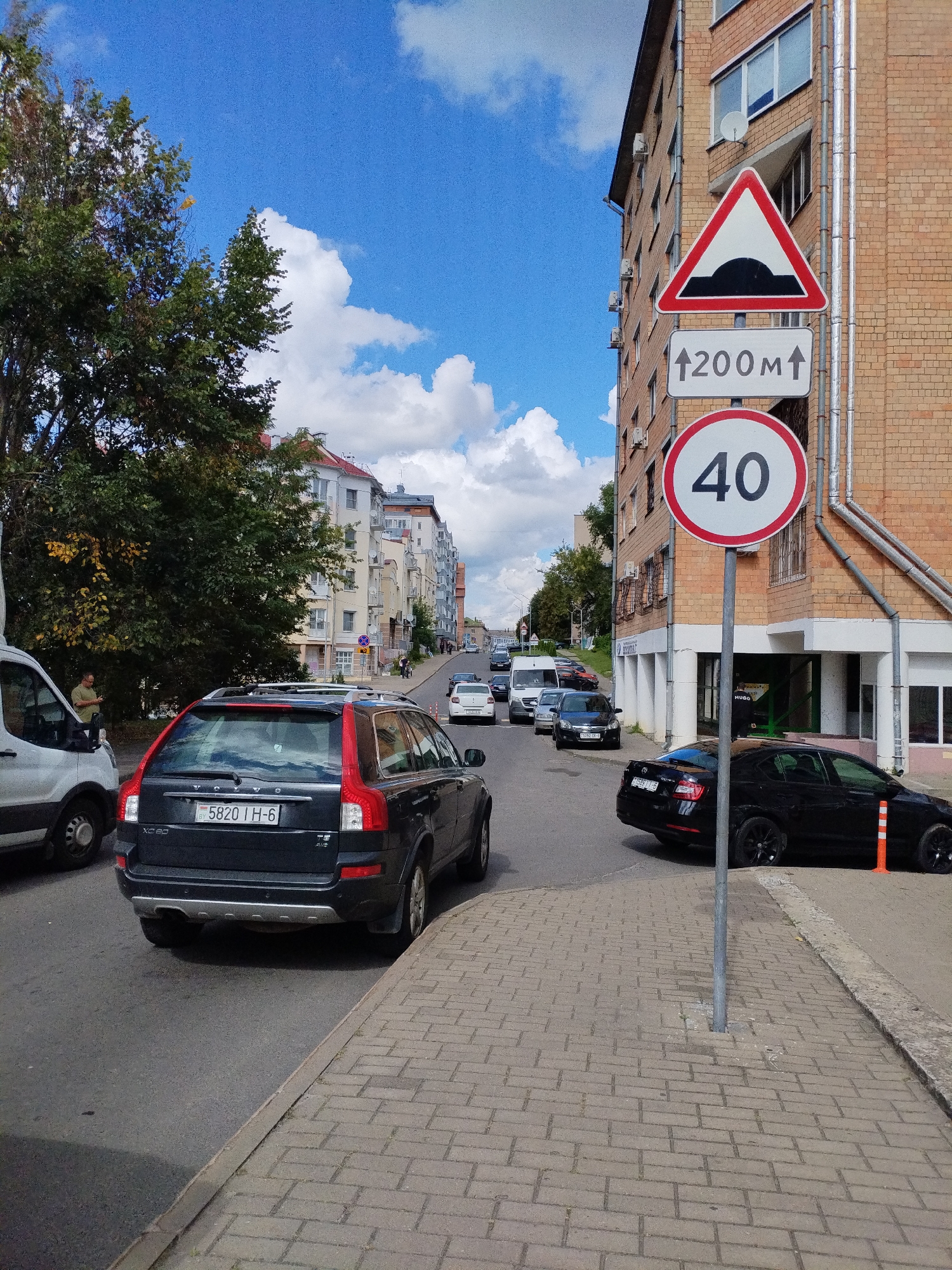 A street scene in Mahilyow, Belarus, under a partly cloudy blue sky. The perspective is from the sidewalk looking down a paved street lined with brick apartment buildings. Traffic signs are prominent: a triangular sign with a black speed bump symbol, a rectangular sign with an upward arrow and "200m", and a circular sign with "40" in black on a white background with a red border. Several cars are visible on the street, including a black Volvo XC90, a black sedan, and a white van. Trees with green and yellowing leaves line the left side of the street. A person in a light-colored shirt stands near the white van, looking at a phone. The sidewalk is paved with light-colored bricks.