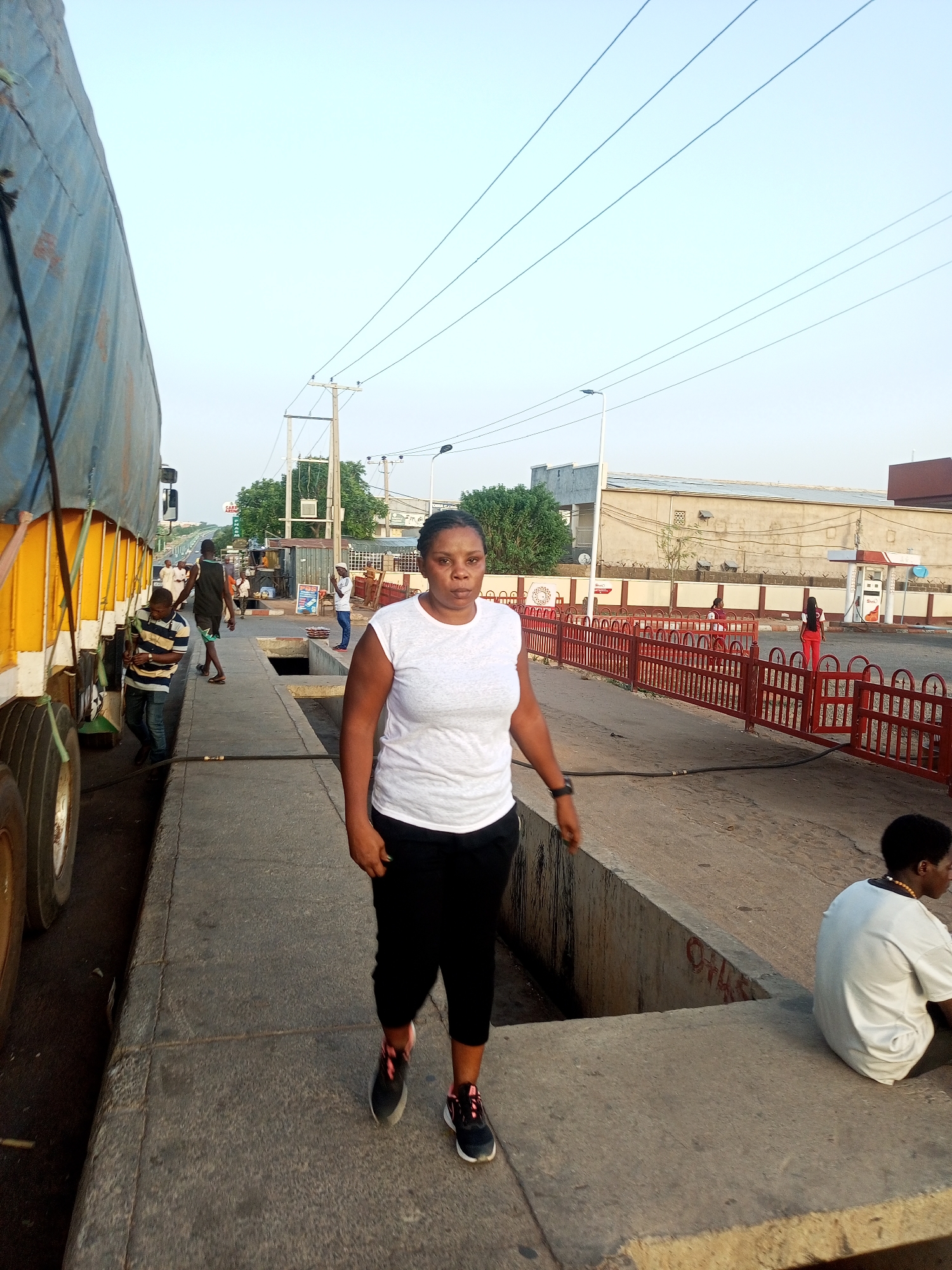 The image depicts a woman walking on a sidewalk in a city street, likely in Unknown, Unknown. The woman is wearing a white t-shirt and black pants with athletic shoes. To the left, a large yellow truck is partially visible, and several people are gathered near it. Behind the woman, there is a concrete barrier with an opening leading to a lower level, where another person is sitting. In the background, there is a street with a red fence, a gas station, and buildings. Overhead, power lines stretch across the sky.