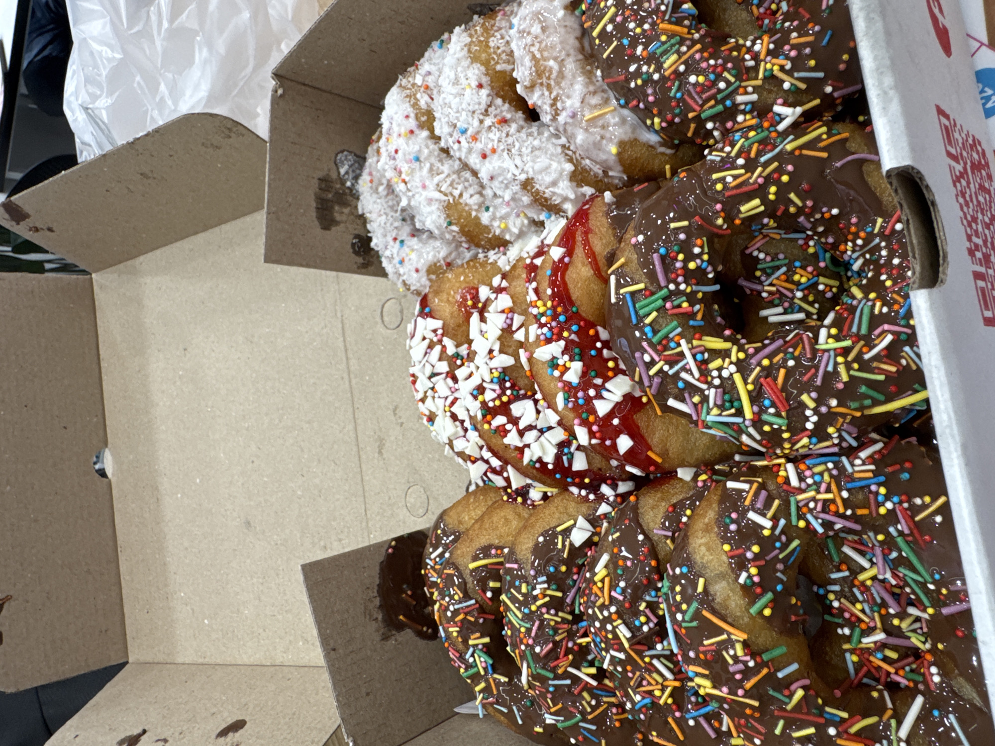 A partially opened cardboard box contains several donuts with various toppings. One donut is covered in shredded coconut and small colorful sprinkles, another has a red glaze with white and colorful sprinkles, and the rest are covered in chocolate frosting and multicolored sprinkles. The box is sitting on a surface with a plastic bag partially visible in the upper left corner. Location is unknown.