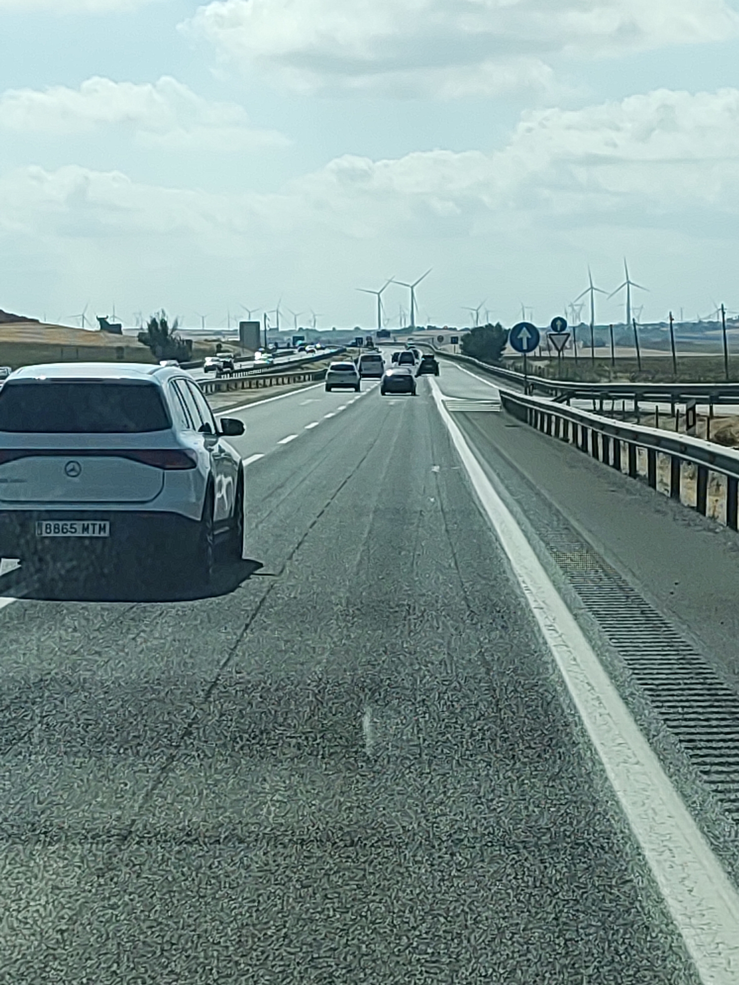 The image shows a highway with several cars on it. In the foreground, there is a white Mercedes-Benz EQE sedan with the license plate "BB65 MTM". The car is in the right lane of a highway. In the background, there are several wind turbines. The sky is cloudy. The location is the AP-2 highway, near Castillo de Garcimuñoz, Spain. The highway has multiple lanes and is bordered by a guardrail and a dry, grassy shoulder. There are road signs visible, including one with a blue circle and a white arrow pointing upwards, indicating a straight-ahead lane. The overall scene depicts a typical highway environment with vehicles moving and infrastructure for renewable energy in the distance.