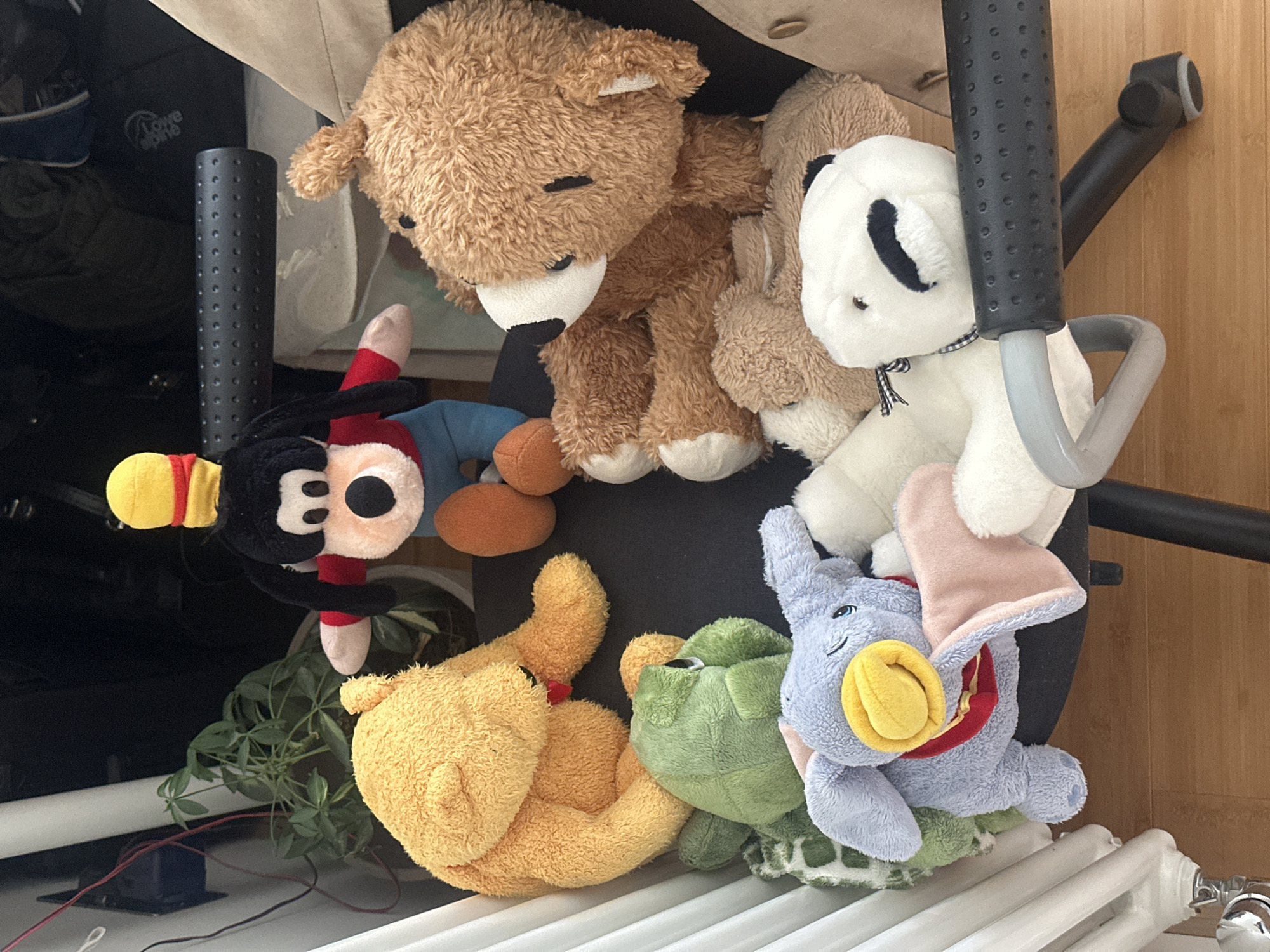 The image depicts a collection of plush toys piled on a white radiator and partially obscured by a black office chair. The toys include a brown bear, a panda, Mickey Mouse, Winnie the Pooh, a green turtle, and a blue elephant wearing a red scarf. The background includes a beige bag, a black object, and a plant. The scene takes place indoors with a wood-paneled wall visible. The location within Wealden, United Kingdom cannot be determined from the provided image.