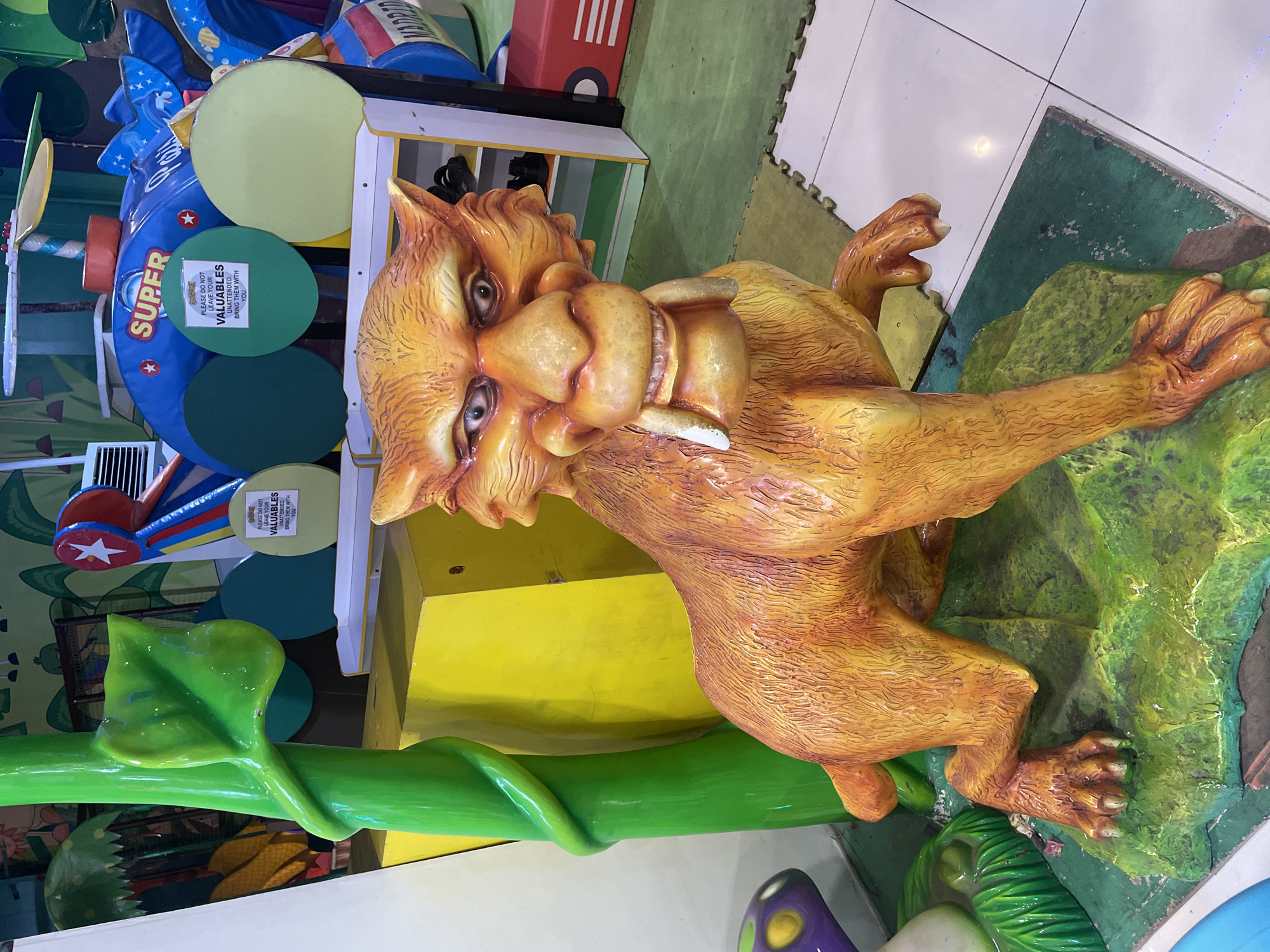 The image shows a brightly colored, life-size statue of a sabre-toothed cat, likely made of fiberglass or plastic, in what appears to be an indoor amusement or play area. The statue is orange with yellow and red accents, and it is positioned on a green, rocky base. Behind the statue, there is a large yellow structure, a green vine-like sculpture, and a blue and red helicopter-themed structure with the word "SUPER" prominently displayed. Several green and yellow circular objects are attached to the helicopter structure. A sign on one of the green circles reads "PLEASE DO NOT LEAVE YOUR VALUABLES UNATTENDED. BRING THEM WITH YOU." The floor is covered with interlocking mats and white tiles. The overall scene suggests a whimsical and child-friendly environment. This location is likely in the city of Davao City, Philippines, as indicated by the context of similar amusement facilities found in the region.
