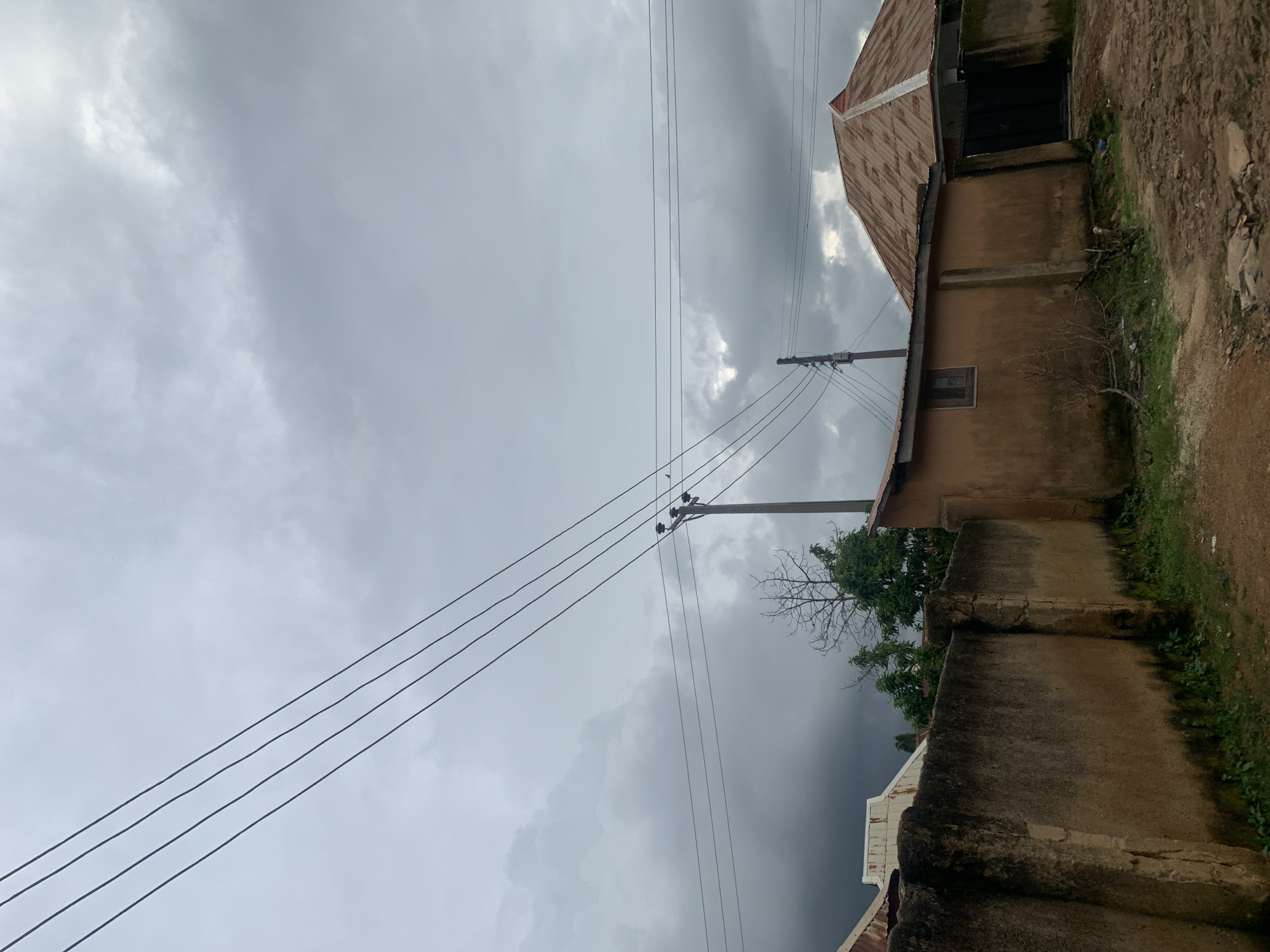 The image depicts an overcast sky with dense, dark clouds gathering, suggesting impending rain over a residential area in Rafin Par, Nigeria. In the foreground, a textured, weathered concrete wall runs horizontally across the frame, appearing aged and partially covered in moss and green vegetation at its base. To the right of the wall, a section of an orange-brown building is visible, featuring a small, recessed window and a corrugated metal roof. Behind this building, another structure with a more complex, multi-faceted corrugated metal roof is partially visible, extending to a dark gate. Multiple black electrical wires stretch diagonally across the sky, supported by a utility pole with insulators situated on the right side of the image. A few sparse, bare tree branches are seen emerging from behind the wall and buildings. The ground appears to be a dirt path with some scattered stones and debris.