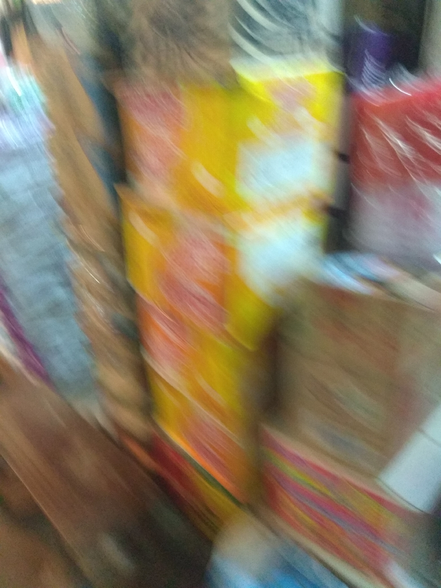 The image is a blurry, abstract depiction of what appears to be a retail display. The dominant colors are yellow, red, and blue, with streaks and blurs suggesting movement. The subjects are indistinct objects, possibly stacked boxes or packages, with some wrapping visible. There are no discernible people or clear signs of a specific event. The lighting is somewhat dim, and there are no obvious cues about the time of day or weather. The motion blur makes it impossible to identify any text, specific location details, or emotions. The overall impression is one of a vibrant, but chaotic, commercial environment, possibly a market stall or shop interior in Jalingo, Nigeria, given the location context.