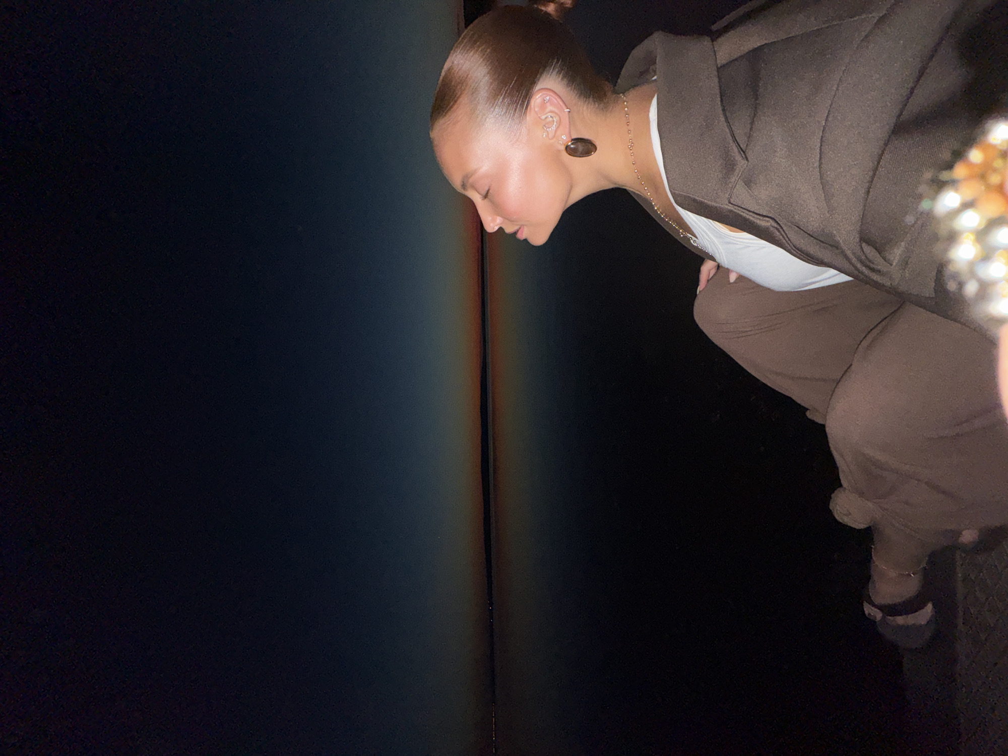 The image captures a woman seated outdoors at dusk or early night, looking upwards with her eyes closed and a serene expression. The scene is located in the city of Chaikovsky, Russia.
The background features a dark sky, transitioning to a band of warm orange and red light along a flat horizon, indicative of either a recent sunset or impending sunrise. A faint, dark landmass with distant lights is visible across a calm, dark body of water that reflects the remaining sky glow.
The woman is positioned in profile, facing right. Her reddish-brown hair is pulled back tightly into a sleek bun. Her left ear is clearly visible and adorned with multiple pieces of jewelry: a prominent, dark, oval-shaped dangle earring, at least three small stud earrings along the earlobe, and a small crescent-shaped earring or cuff higher on the helix. She wears a delicate, gold-toned beaded necklace. Her attire consists of a white top worn under a dark brown, oversized jacket or blazer with a wide collar. She is also wearing loose-fitting, dark brown trousers that match the jacket. Her left hand rests on her thigh, and her left foot, wearing a dark sandal, is visible on a dark, textured ground surface. A blurry, multi-faceted, reflective object, possibly a bracelet, is partially visible in the extreme lower right foreground.
