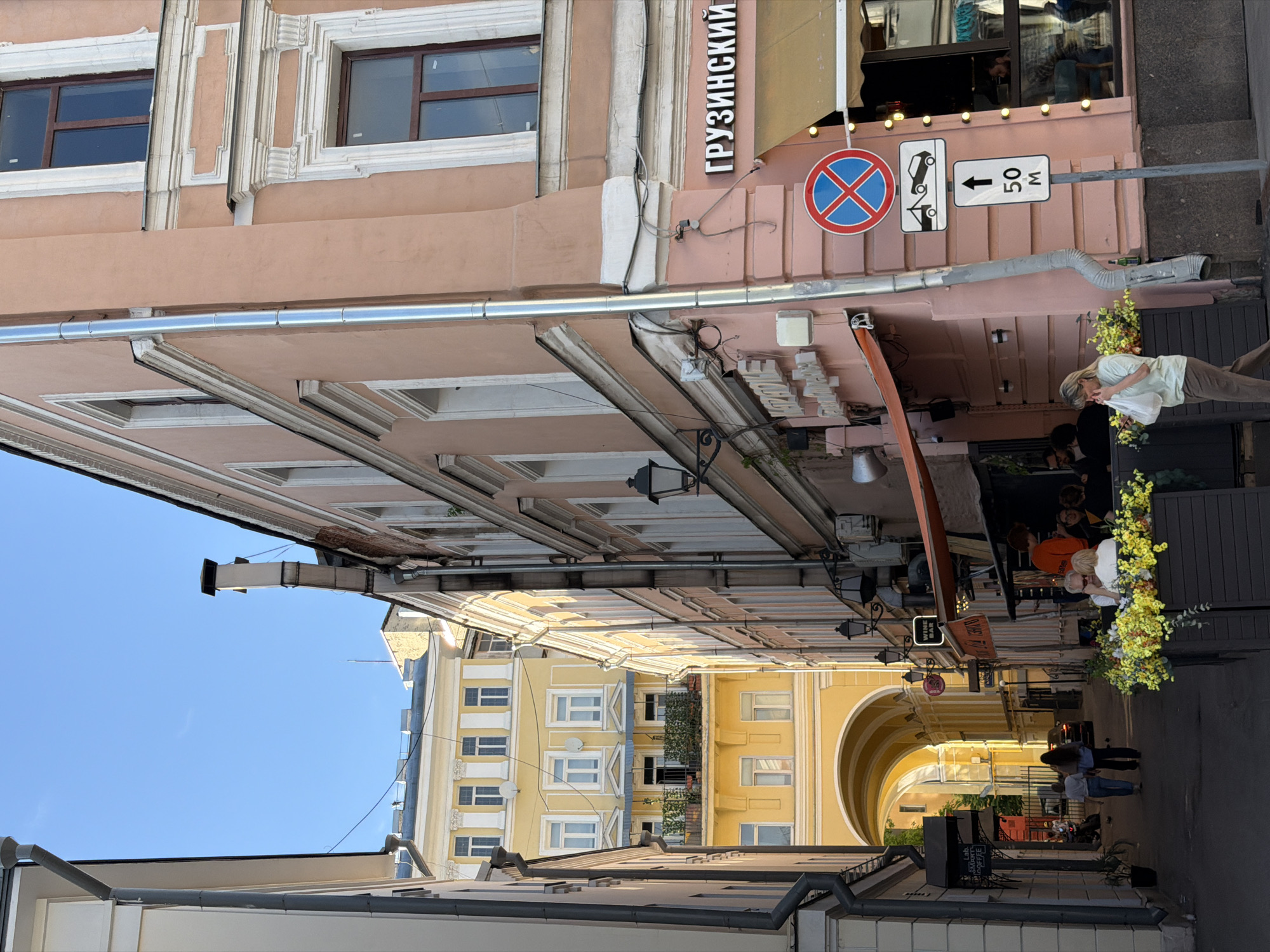 A narrow Moscow alleyway is shown in an eye-level, rotated view, where buildings line the sides. At the end of the alleyway, an arched building connects the structures. There are two stop signs with text that appears to be in the Russian language, as well as an additional sign indicating the distance to the next street. People are walking near the restaurant, and some are seated near flower boxes. The restaurant has an orange awning.
