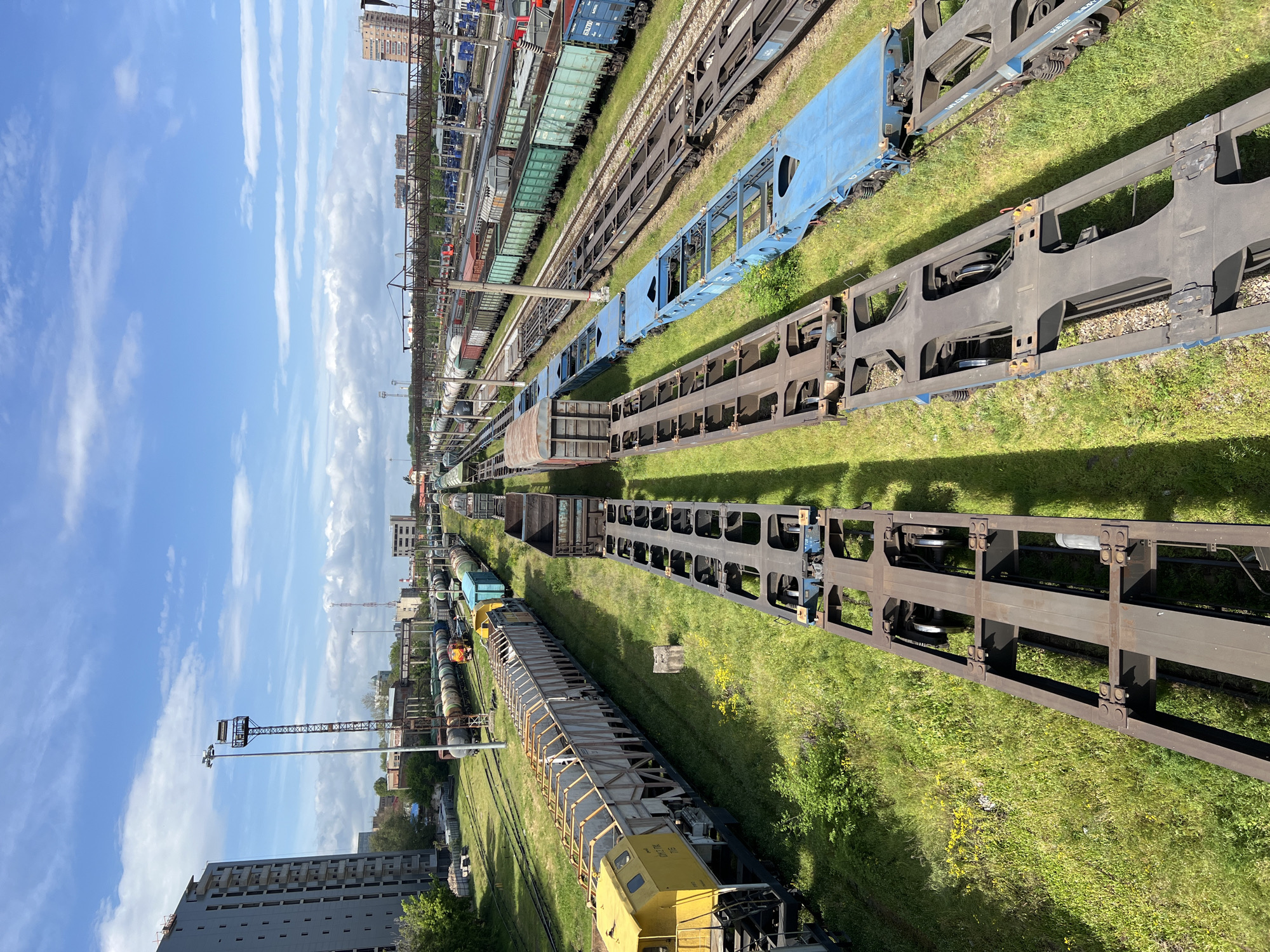 The image depicts a railway yard in Krasnodar, Russia. Multiple parallel tracks are visible, occupied by various types of freight cars. Some of the cars are flatbeds, while others are boxcars. The tracks are surrounded by green grass. A large building is visible in the distance, as well as a light tower. The sky is blue with scattered clouds.