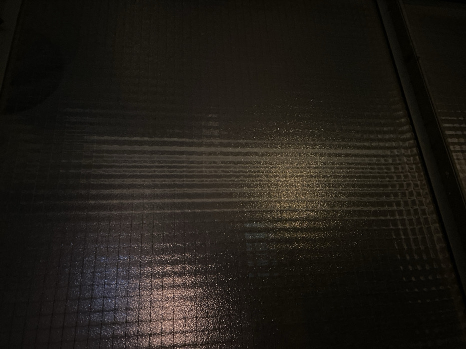 The image displays a dark, rectangular panel with a textured, semi-gloss finish, possibly indicative of a wall or a large door. The texture forms a subtle grid-like pattern across the surface. Prominent vertical streaks of a lighter, off-white hue are visible, concentrated on the left and central portions of the panel, consistent with dried liquid residue or water flow marks. The surface exhibits reflective properties, showing blurred, diffused light sources. A reddish-orange light reflection is present in the upper-left quadrant, while a warmer, yellowish-white light reflection is visible in the lower-central left. A distinct horizontal black band or seam runs across the bottom edge of the frame. No individuals, specific objects, or actions are depicted. The scene suggests an interior or sheltered exterior urban environment, likely in low light conditions, such as within the city of Tel-Aviv, Israel, although no specific landmarks or precise location details are discernible from the image itself.