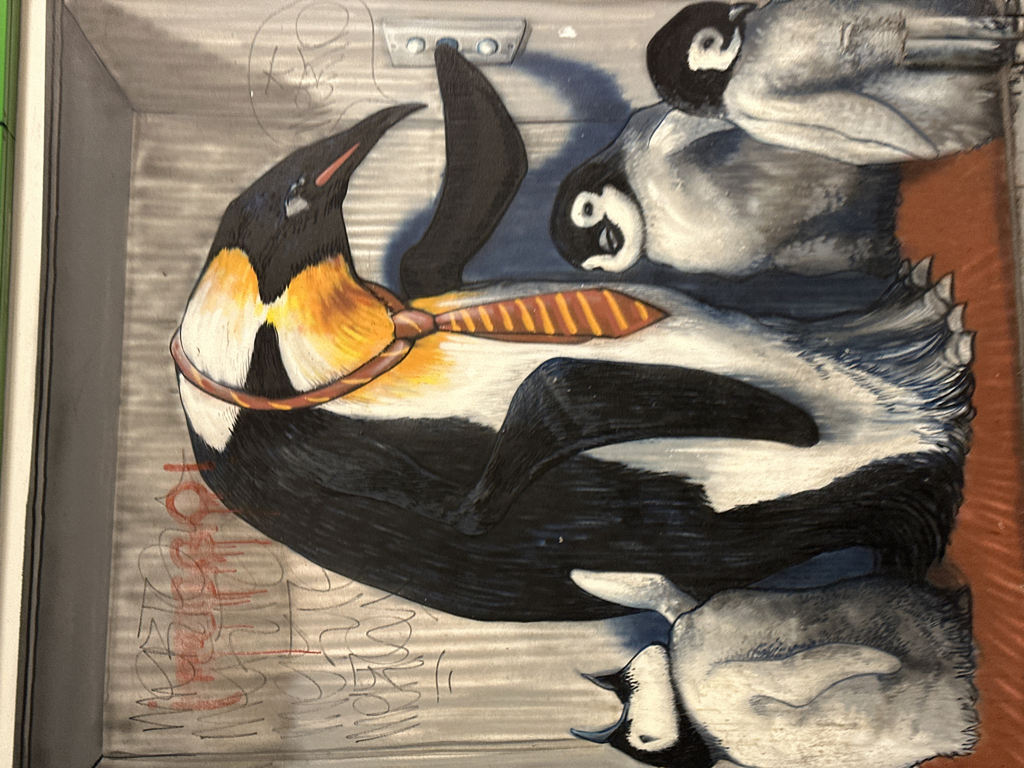 The image shows a piece of street art, potentially graffiti, depicting an emperor penguin wearing a tie. The penguin is surrounded by three penguin chicks. The artwork appears to be painted on a textured surface, perhaps a shuttered metal door or similar urban fixture. There are additional markings or graffiti around the central penguin image, including what appears to be red lettering. The setting is suggested to be Milan, Italy.