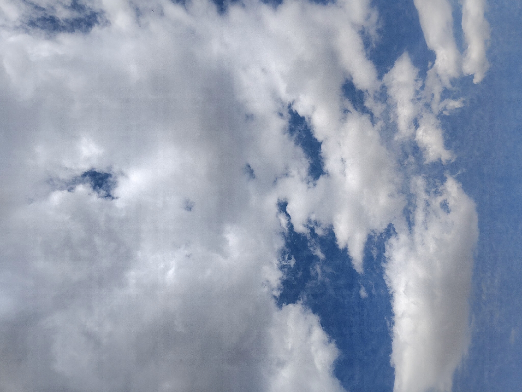 The image shows a sky with scattered clouds over a landscape that is not visible. The sky is a mix of blue and white clouds, with some darker, thicker clouds and some lighter, thinner clouds. There are areas of clear blue sky visible between the clouds. The overall impression is of a partly cloudy day. There are no people, subjects, or objects other than the clouds and sky visible in the image. The location in Kopuro Bazar, Kyrgyzstan is not discernible from the image itself.