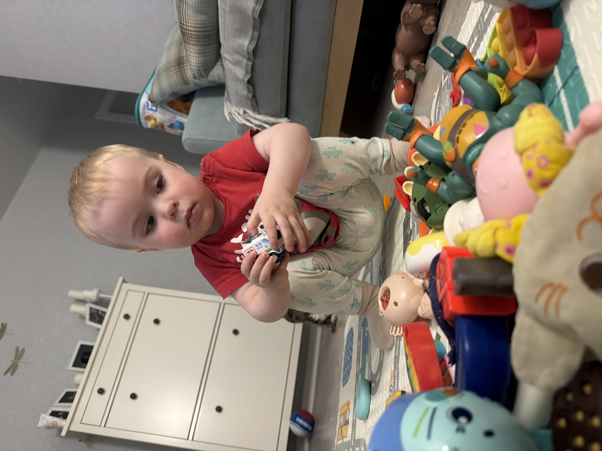 A young child with fair skin and blond hair, wearing a red shirt and patterned pants, is seated on a patterned play mat. The child is holding a small toy car. The scene is indoors, with a white dresser visible in the background, featuring small decorative objects and framed images on top. Toys are scattered on the play mat, including colorful plastic objects and plush animals. A portion of a couch with a plaid pillow is visible in the background. There is no information about the location being in Votkinsk, Russia.