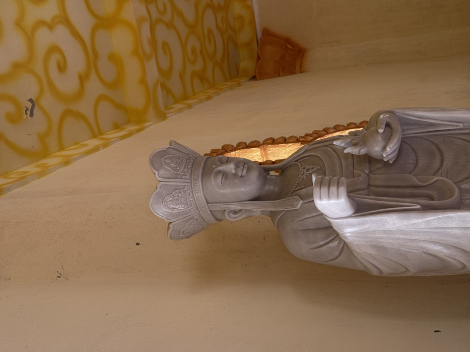This image presents a light-colored stone statue of a crowned figure situated within an interior architectural space in Đăk Mar, Vietnam. The statue displays a serene facial expression, an elaborate multi-tiered necklace, and draped robes. The figure's proper right hand is raised, holding a small, indistinct object or performing a gesture, while the proper left hand cradles a spherical object topped with a flame-like finial. The immediate environment includes plain, light-yellow colored walls. To the proper left of the statue, a narrow, vertical opening with a ribbed frame emits a warm, orange-yellow light. The ceiling is white, decorated with extensive yellow, curvilinear cloud patterns. A decorative architectural bracket, colored yellow, is located at the junction of the wall and ceiling in the upper proper left section of the frame.
