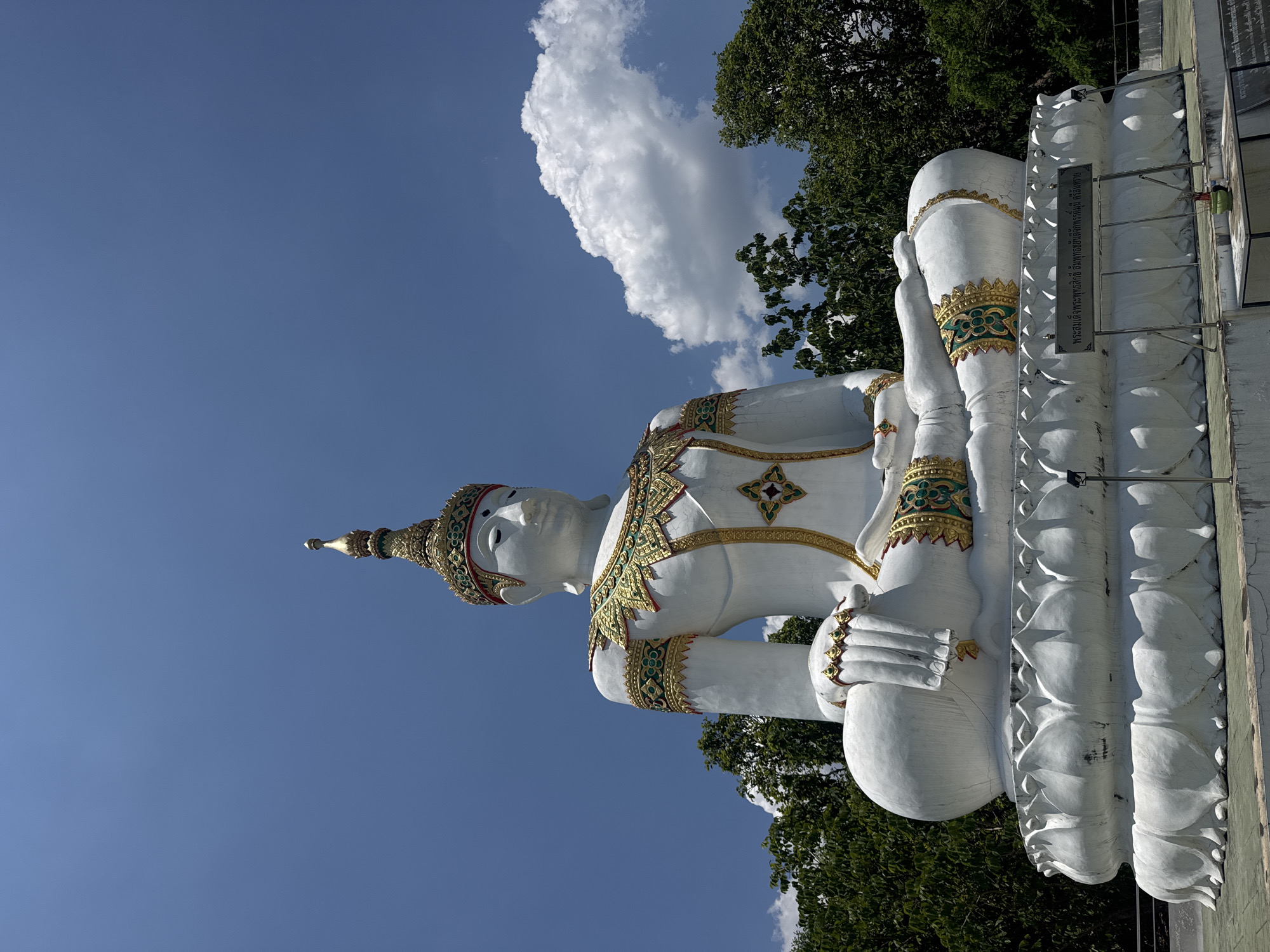The image presents a large, white statue in the lotus position, ornamented with golden and green details. The statue is set against a backdrop of a blue sky with a fluffy white cloud to the left and green foliage from trees above and below. The statue appears to be seated on a detailed base with lotus motifs and a sign in Thai script. The statue is located in Nong Yaeng, Thailand.