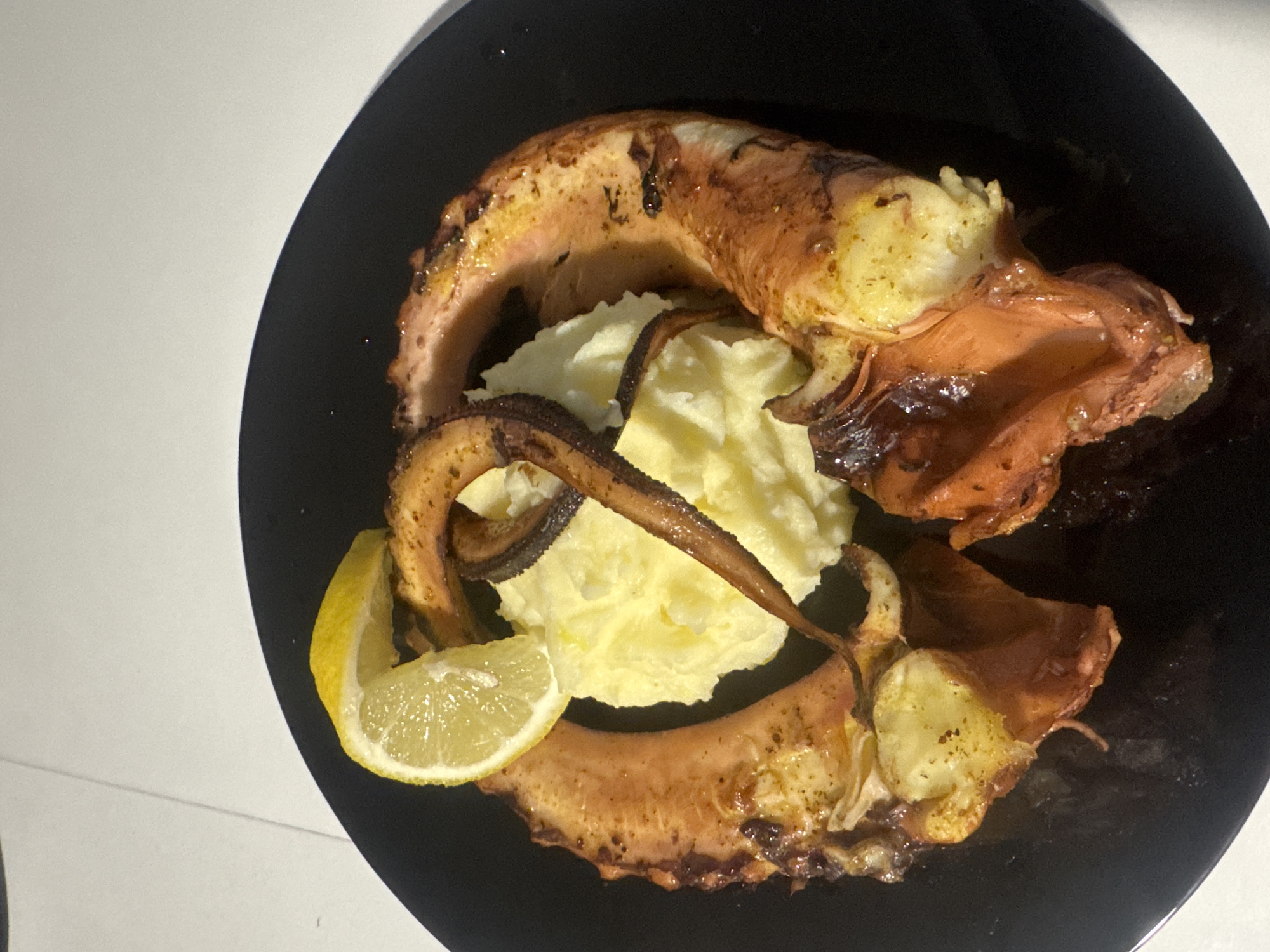 A close-up, elevated shot displays a single serving of food on a black, round plate. The main subject consists of large, cooked octopus tentacles, exhibiting a reddish-brown, somewhat charred surface, arranged in a curved pattern. Centered within the octopus is a mound of mashed potatoes, light yellow in color. A single bright yellow lemon slice rests atop one of the octopus tentacles, partially covering the mashed potatoes. The plate is situated on a light-colored, possibly white or off-white, surface that shows faint lines or seams. There are no individuals, actions, or interactions depicted. This scene is located in Varna, Bulgaria.